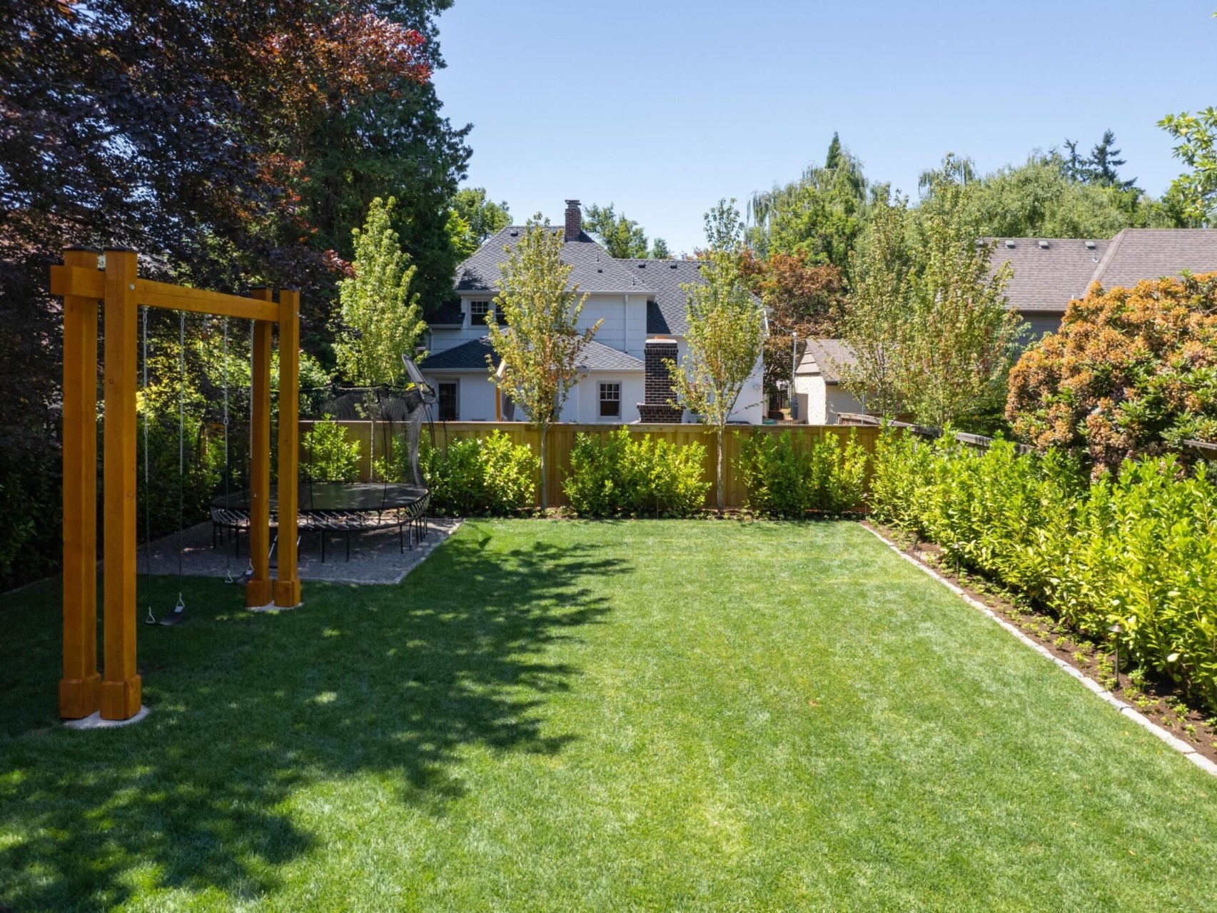 A well-maintained backyard in Portland, Oregon, with lush green grass is surrounded by a wooden fence and various trees. A wooden pergola stands on the left, and a trampoline is visible. Houses with slanted roofs can be seen in the background under a clear blue sky—a dream for any Portland Realtor.