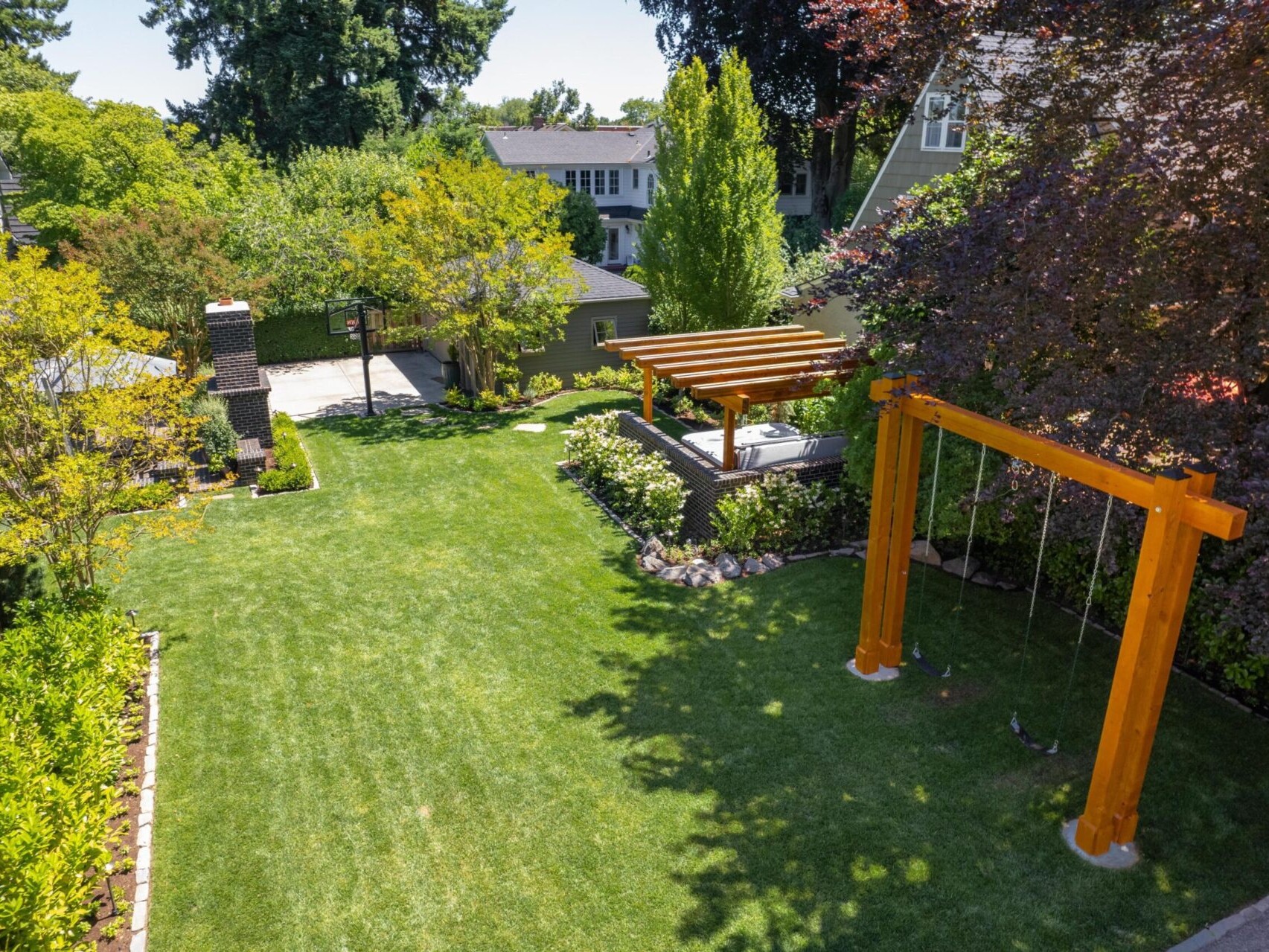 A lush backyard in Portland showcases a green lawn, trees, and a wooden swing set. A pergola-covered seating area is surrounded by vibrant plants. A stone chimney stands against houses and tall trees—an idyllic scene that any top real estate agent would highlight in Portland Oregon real estate listings.