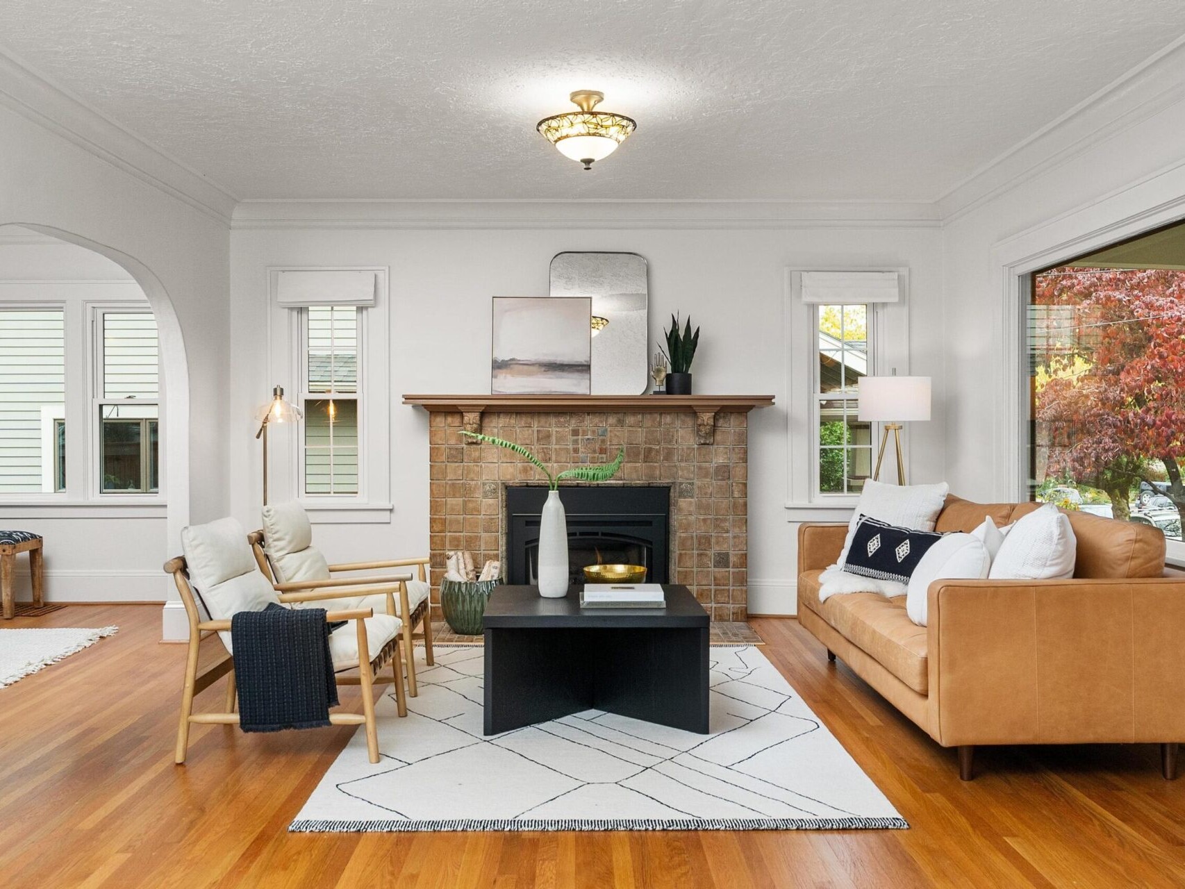 A cozy living room with a brown leather sofa, two white cushioned chairs, a black coffee table, and a fireplace centered on the back wall. The floor is hardwood, and large windows let in natural light. The decor is minimal and modern.