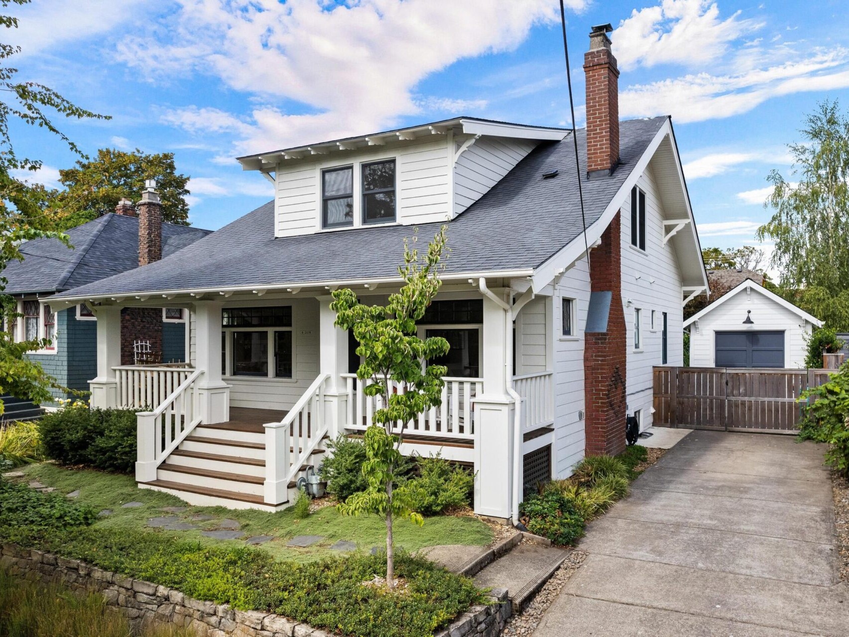 A charming white two-story house in Portland, Oregon, with a covered front porch surrounded by greenery. It features a stone path leading to the steps, a long driveway, and a detached garage. The partly cloudy sky adds to the serene atmosphere.