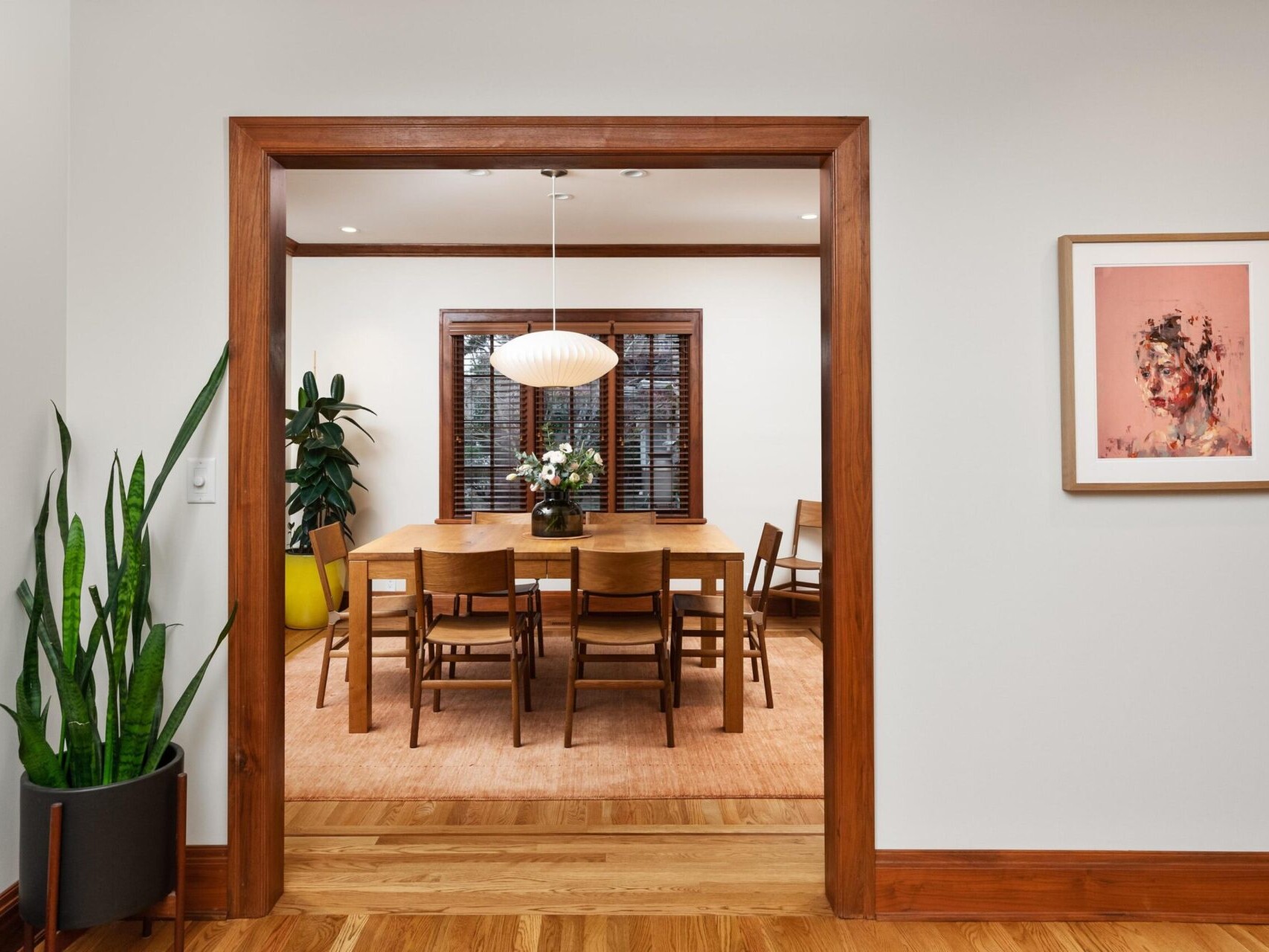 A dining room features a wooden table with eight chairs under a pendant light, perfectly capturing Portland Oregon real estate charm. A large plant graces the left, while framed artwork adorns the right wall—all viewed through a wooden doorway. Ideal for showcasing by a top real estate agent.
