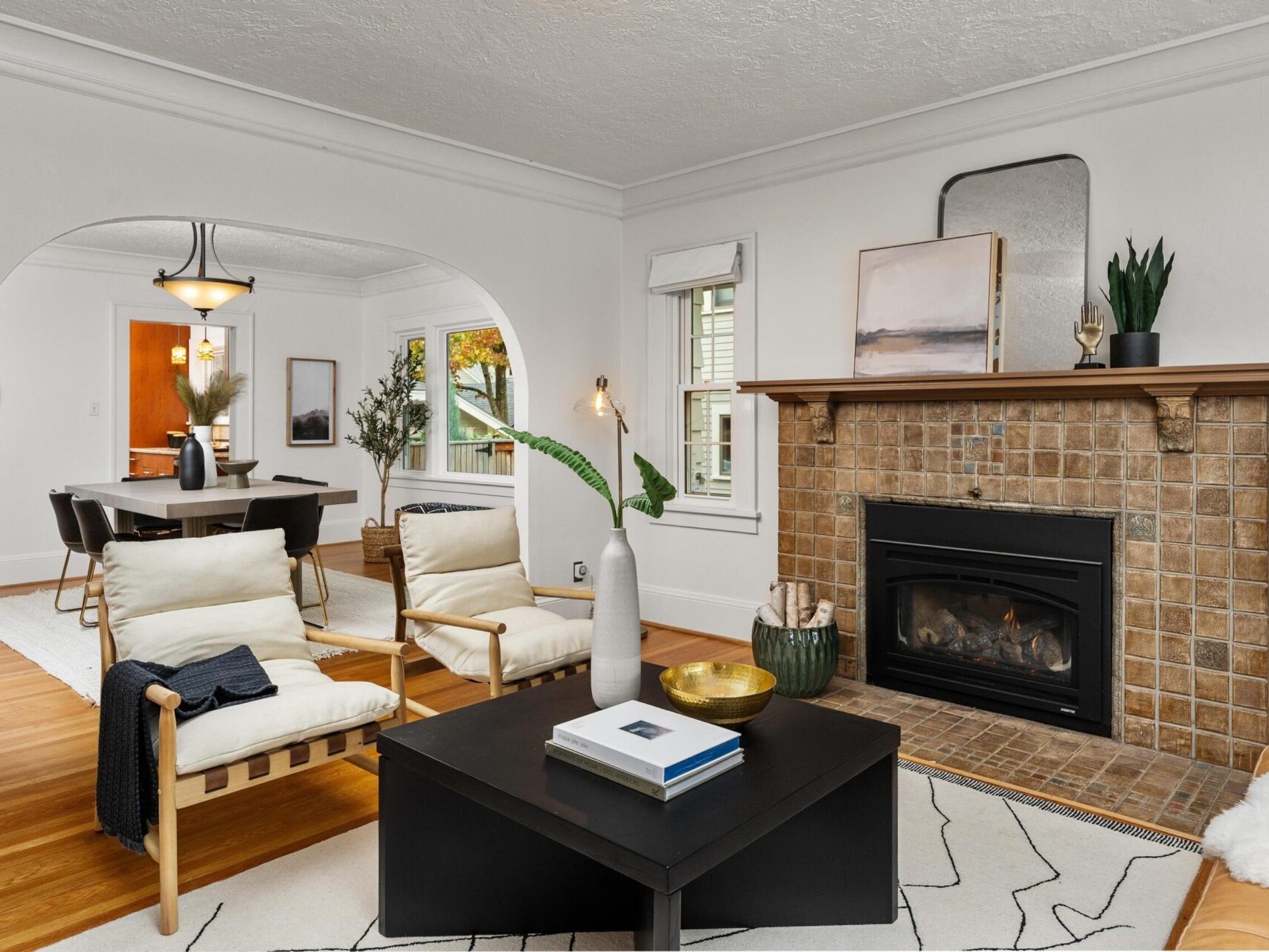 A cozy living room with a tiled fireplace at the center. White walls and wooden floors complement modern furniture, including armchairs and a coffee table. A dining area is visible through an arched opening, featuring a table with chairs.