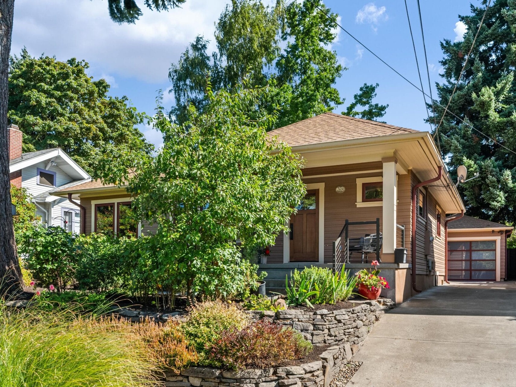 A cozy brown house in Portland, Oregon features a front porch surrounded by lush greenery and a stone wall. The driveway leads to a garage in the background, with tall trees framing the scene under a partly cloudy sky.
