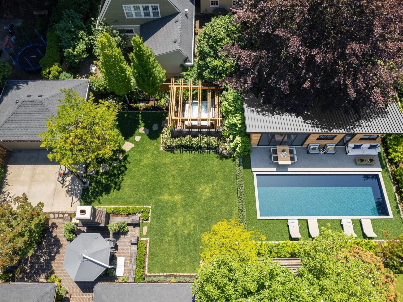 Aerial view of a backyard in Portland, Oregon, featuring a rectangular swimming pool surrounded by lush greenery. A modern pool house, pergola, and patio with furniture enhance the property. A separate garage and trampoline complete this gem represented by a top real estate agent.