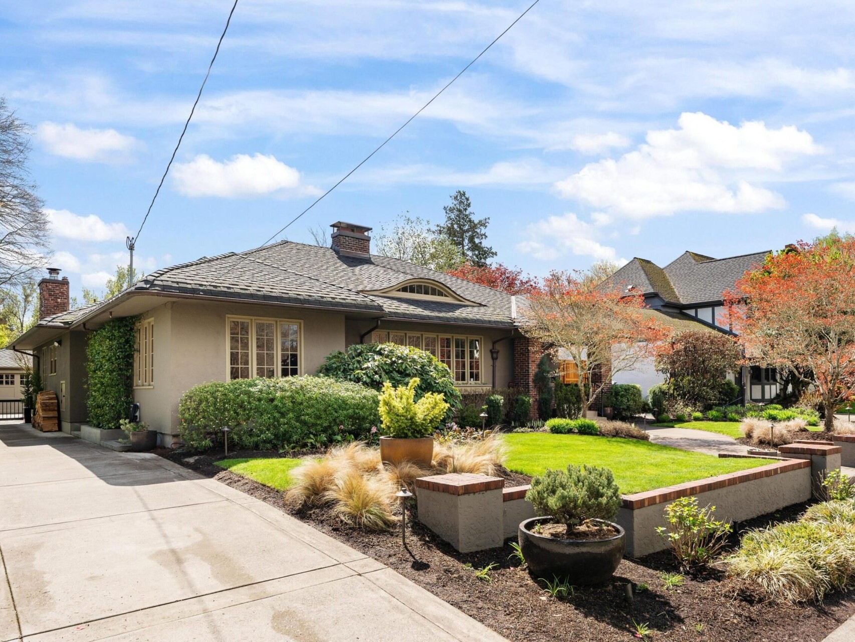 A charming single-story house in Portland, Oregon, with a well-kept lawn and garden. The home features a gray roof, brick and stucco exterior, large windows, and a driveway. Surrounded by trees with red and green foliage under a partly cloudy sky—perfect for Portland real estate enthusiasts.