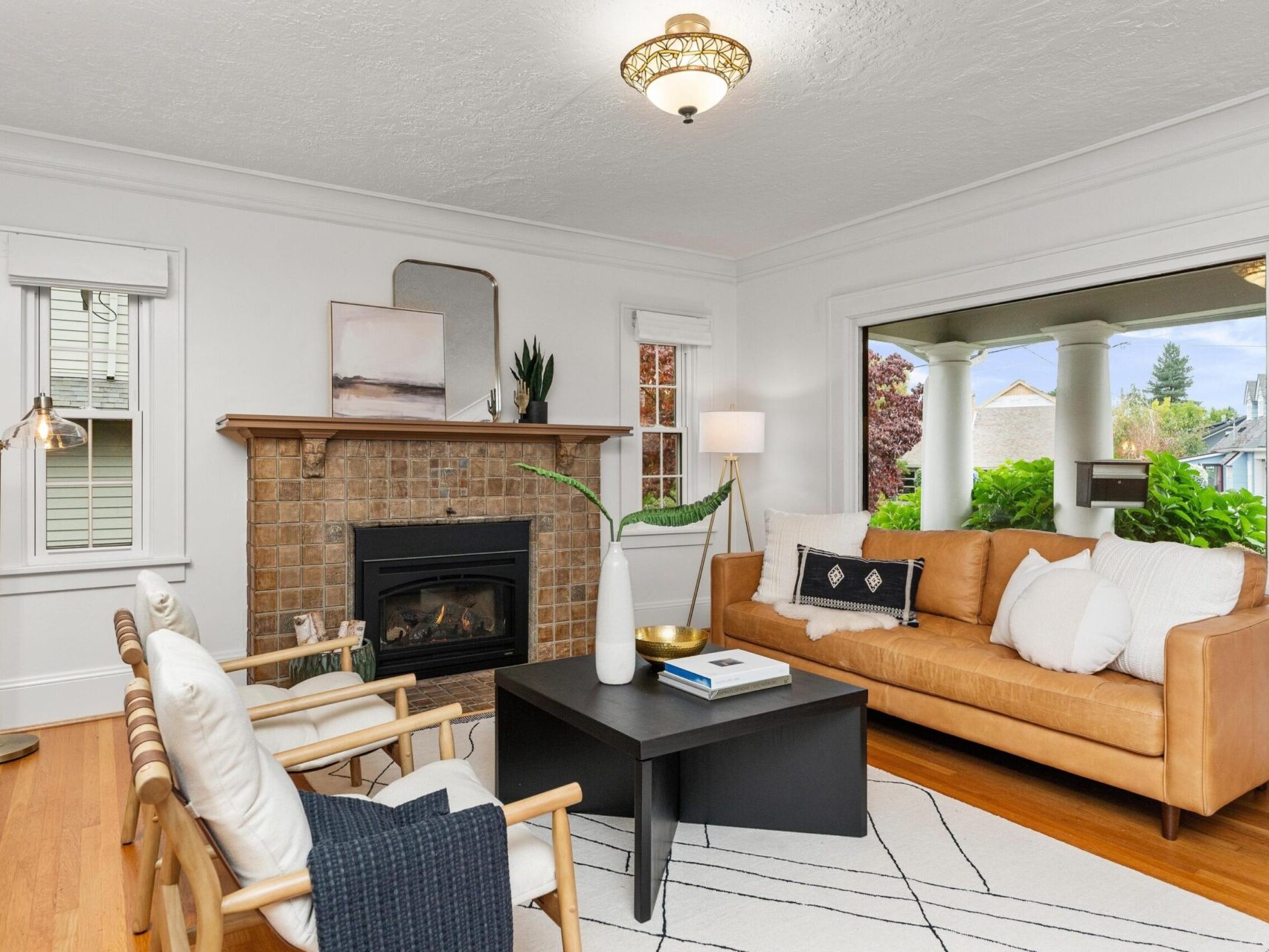 A cozy living room with a tan leather sofa, two white chairs, and a black coffee table on a light geometric rug. A brick fireplace with decorative items, including a painting and plants, sits against a white wall. Large window with a view of greenery outside.