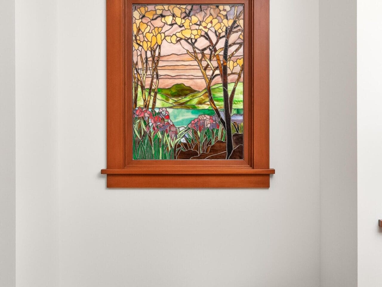 A minimalist Portland, Oregon room features a wooden bench with a folded white blanket. A vibrant stained glass window depicting a landscape with trees, hills, and a colorful sky is framed in rich wood on the wall.