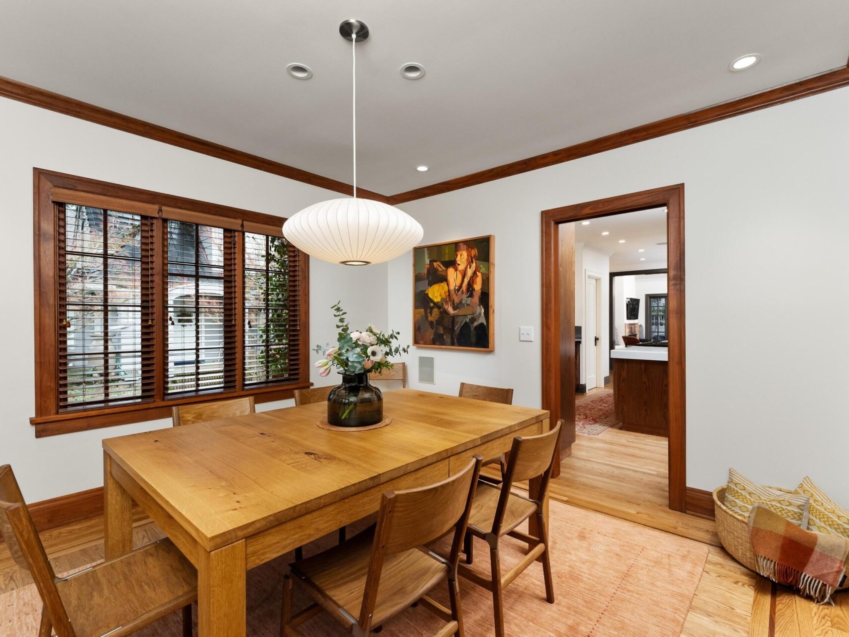 A cozy dining room in a Portland Oregon real estate gem features a wooden table with six chairs on a light rug. A pendant lamp illuminates the space, while flowers in a vase add charm. A large window with blinds invites natural light, and a painting adorns the wall. A hallway leads to another room.