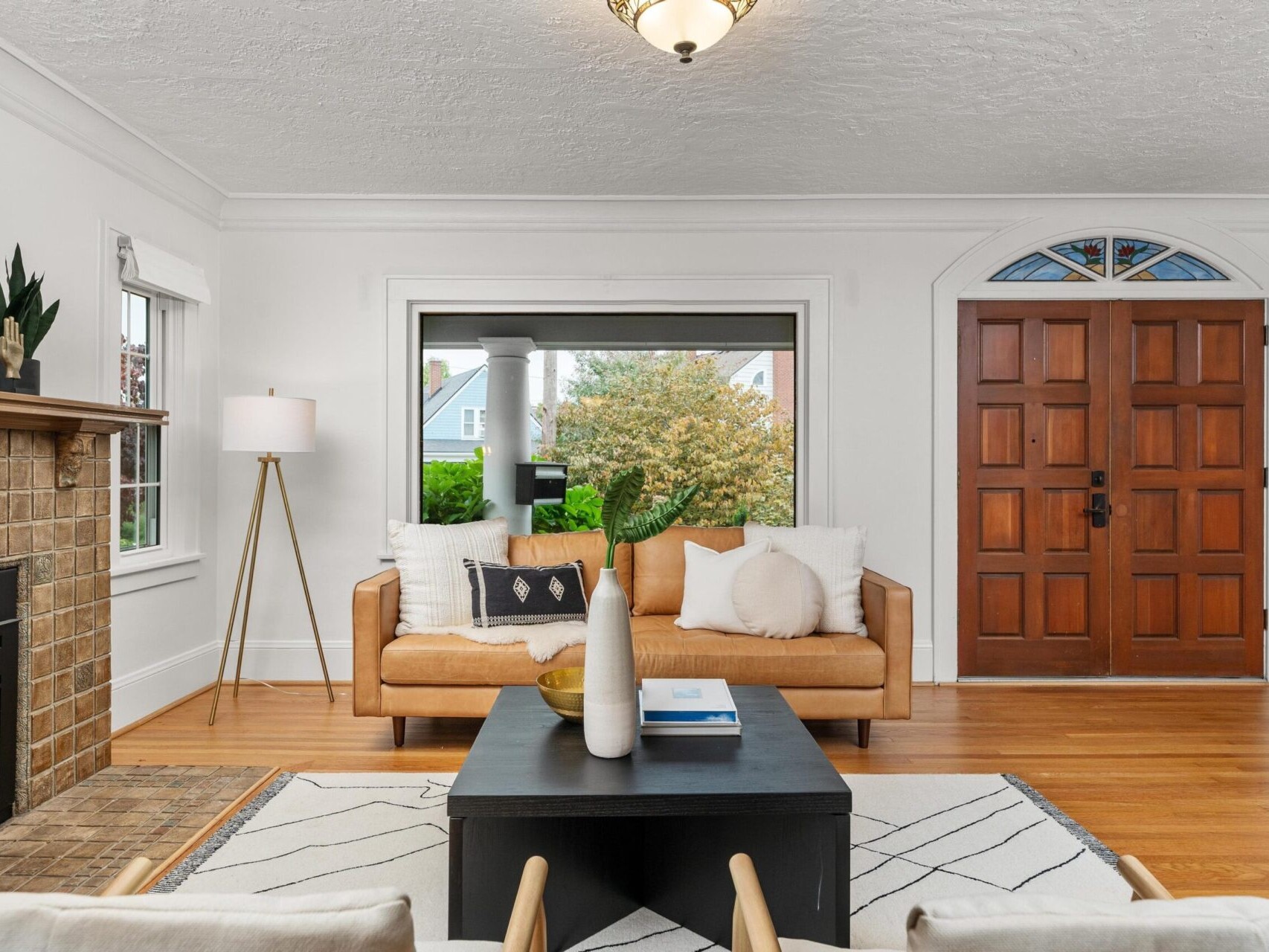 A cozy living room with a tan sofa, black coffee table, and fireplace. Large window with a view of greenery and houses outside. Wooden front door with a decorative glass panel. White walls and hardwood floors complete the elegant space.