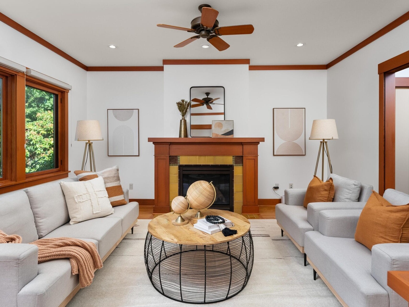 A cozy living room in Portland, Oregon, features two beige sofas facing each other with a round coffee table in between and two lamps on either side of the fireplace. The walls boast abstract art, the ceiling has a fan, and large windows flood the space with natural light.