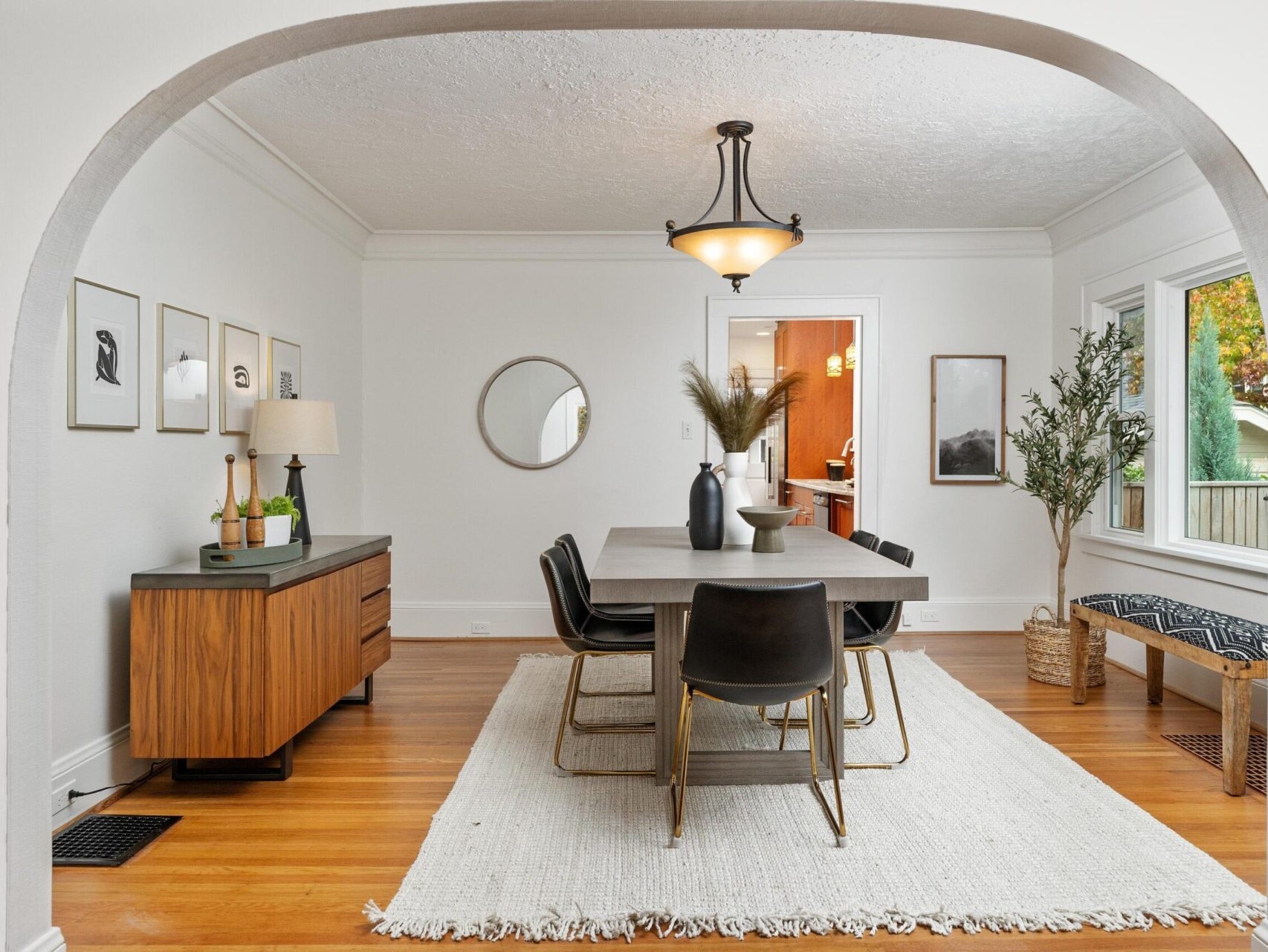 A modern dining room with a wooden table and six black chairs on a white rug. A round mirror and framed art hang on white walls. A sideboard with a lamp and decor is on the left. Large windows and a pendant light provide ample lighting.