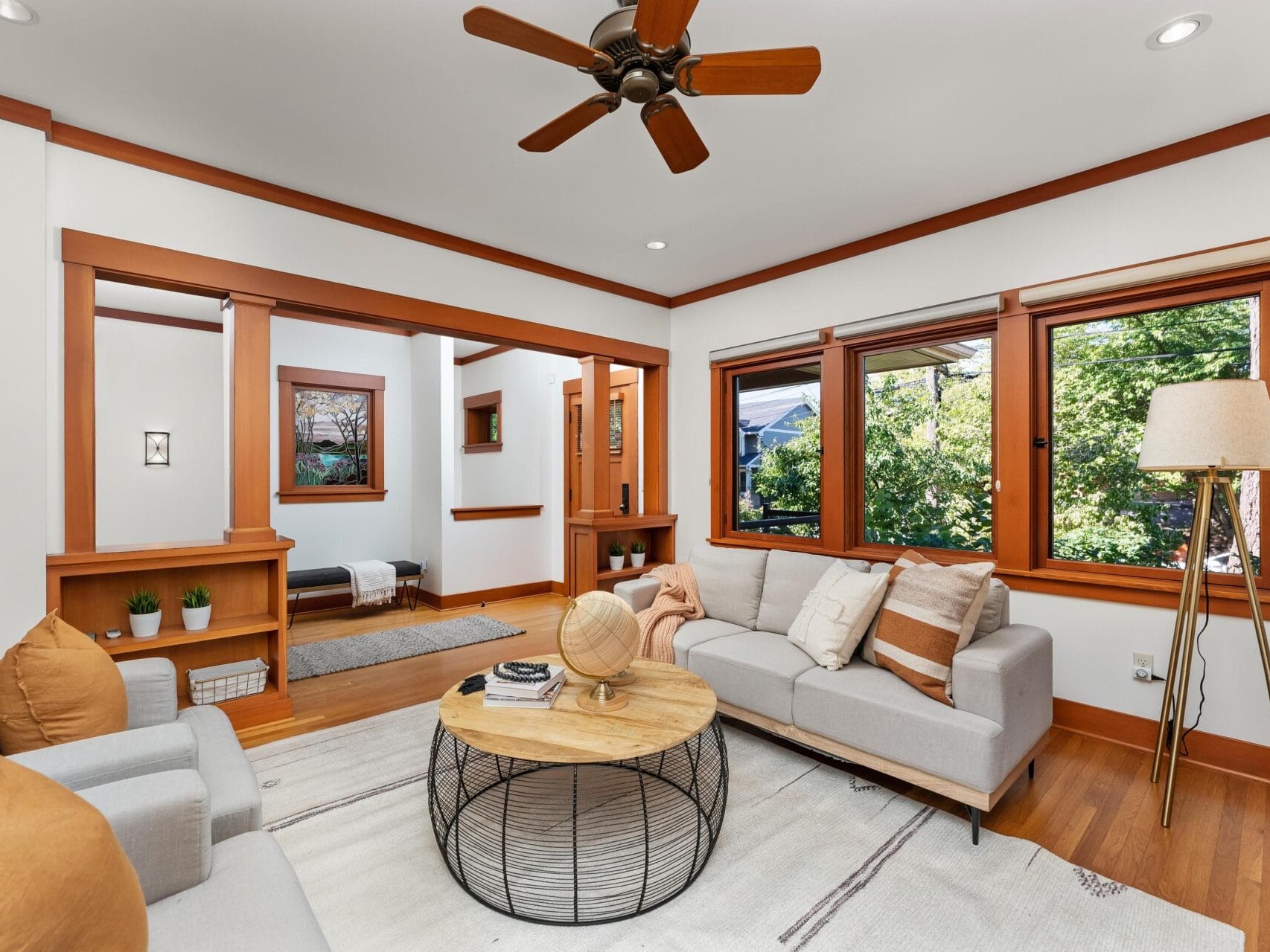A cozy living room in Portland, Oregon, features a light gray sectional sofa with beige and white pillows, a round wooden coffee table, and a floor lamp. Large windows offer natural light and a view of greenery, while a ceiling fan hangs elegantly above.