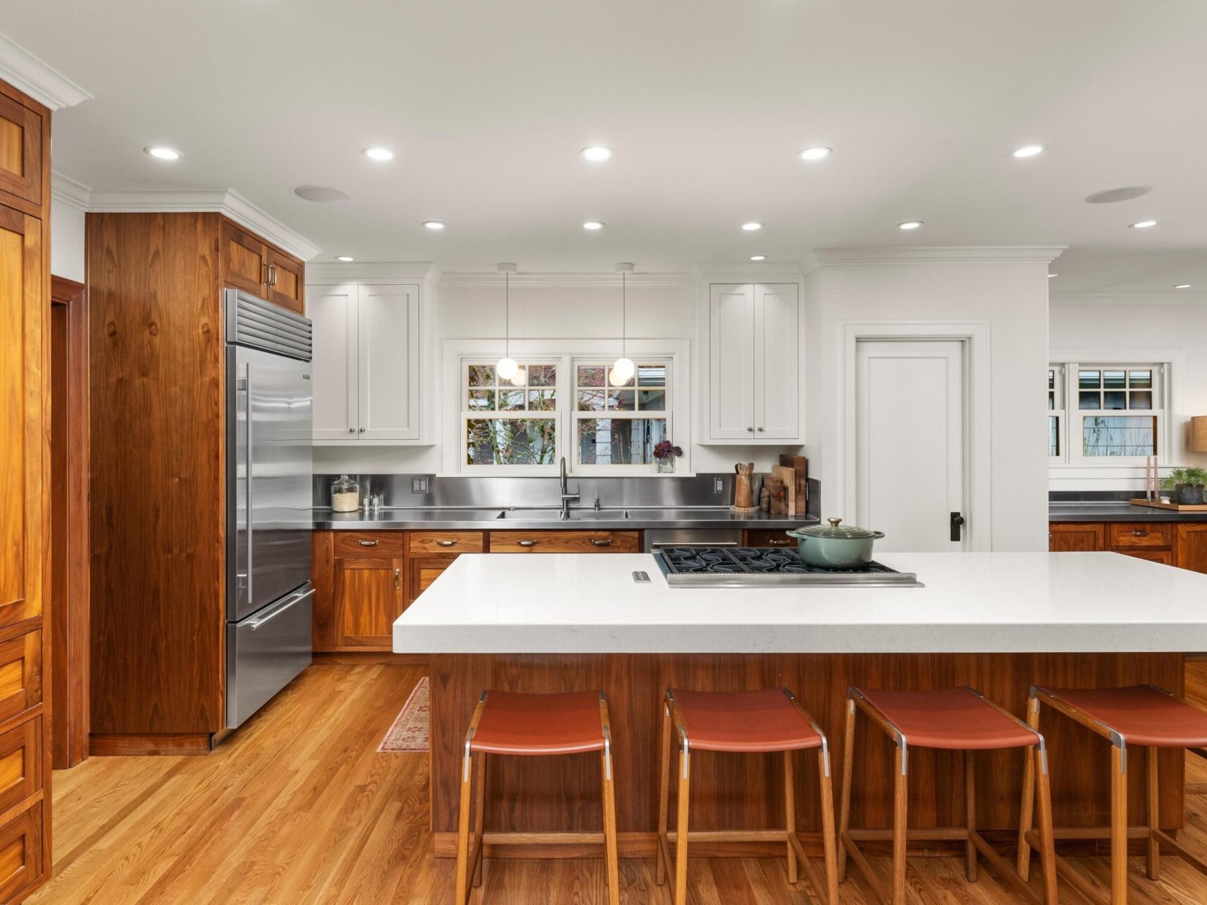 A spacious kitchen with wooden cabinets and a large white island. Four brown stools line the island. Stainless steel appliances, including a fridge and oven, complement the modern design. Perfect for those exploring Portland Oregon real estate, this home shines under recessed lighting.