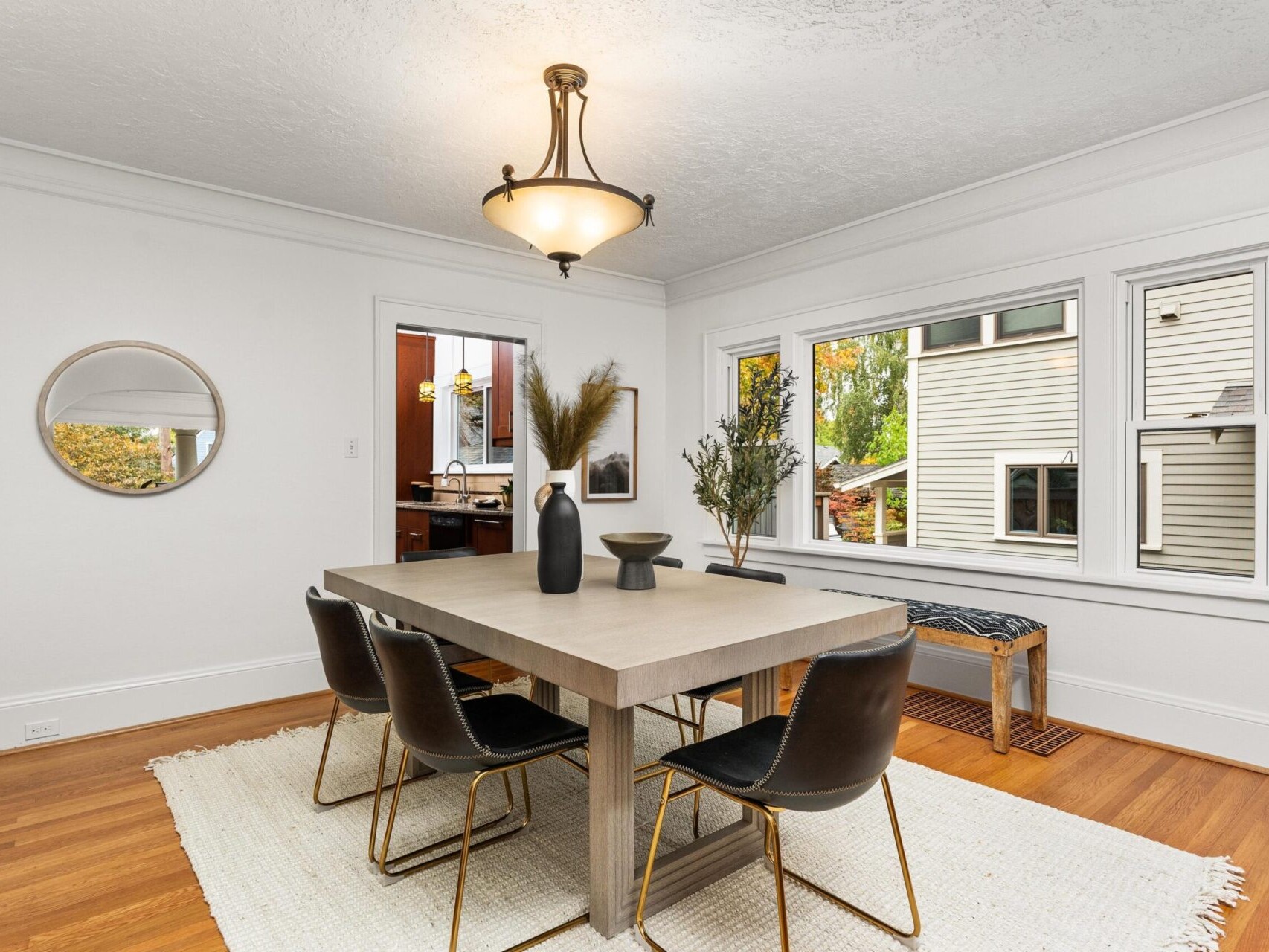 A modern dining room with a rectangular wooden table and six black chairs. The table is decorated with black vases and pampas grass. A round mirror hangs on the white wall, and large windows overlook a house and trees. A beige rug covers the floor.