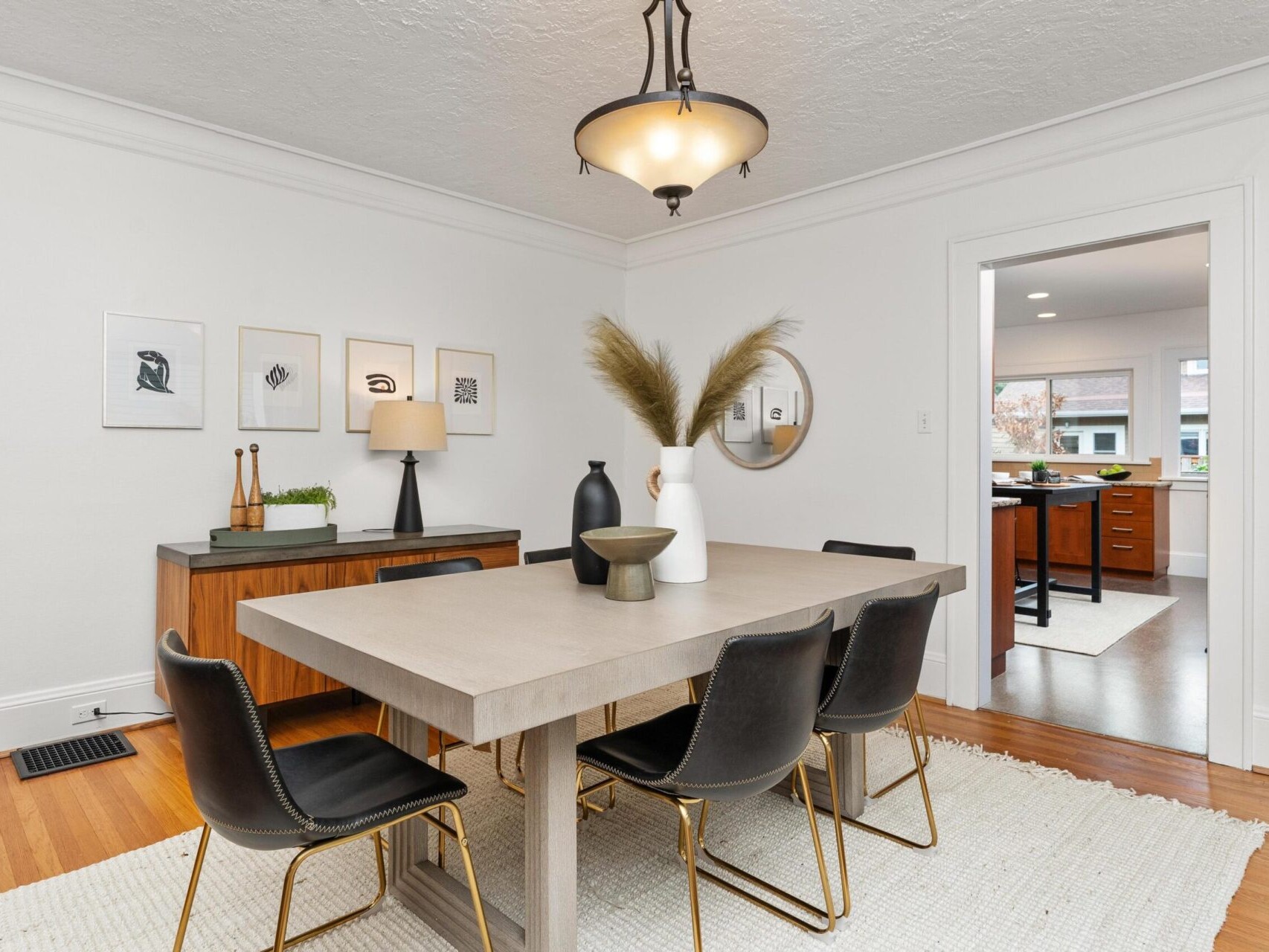 A modern dining room with a concrete table, surrounded by six black chairs. A large vase with pampas grass is on the table. The walls are adorned with minimalist art. A door leads to a kitchen with visible cabinets and a window view.