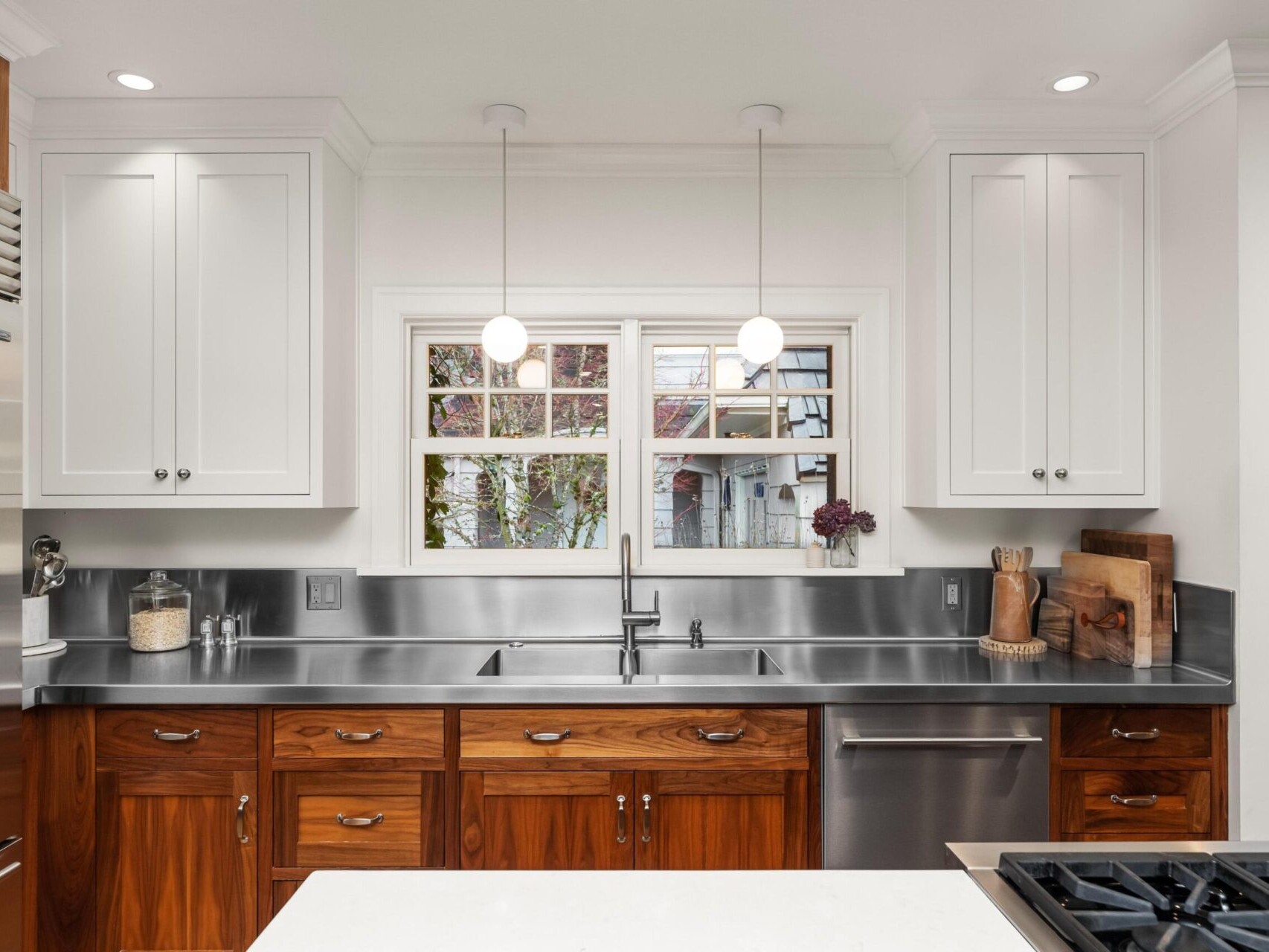 A modern kitchen in Portland Oregon Real Estate, featuring a stainless steel countertop and sink, wooden cabinets, and white upper cupboards. Two pendant lights hang above. A window above the sink offers a view outside. Kitchen items are neatly arranged on the counter for your dream home setup.
