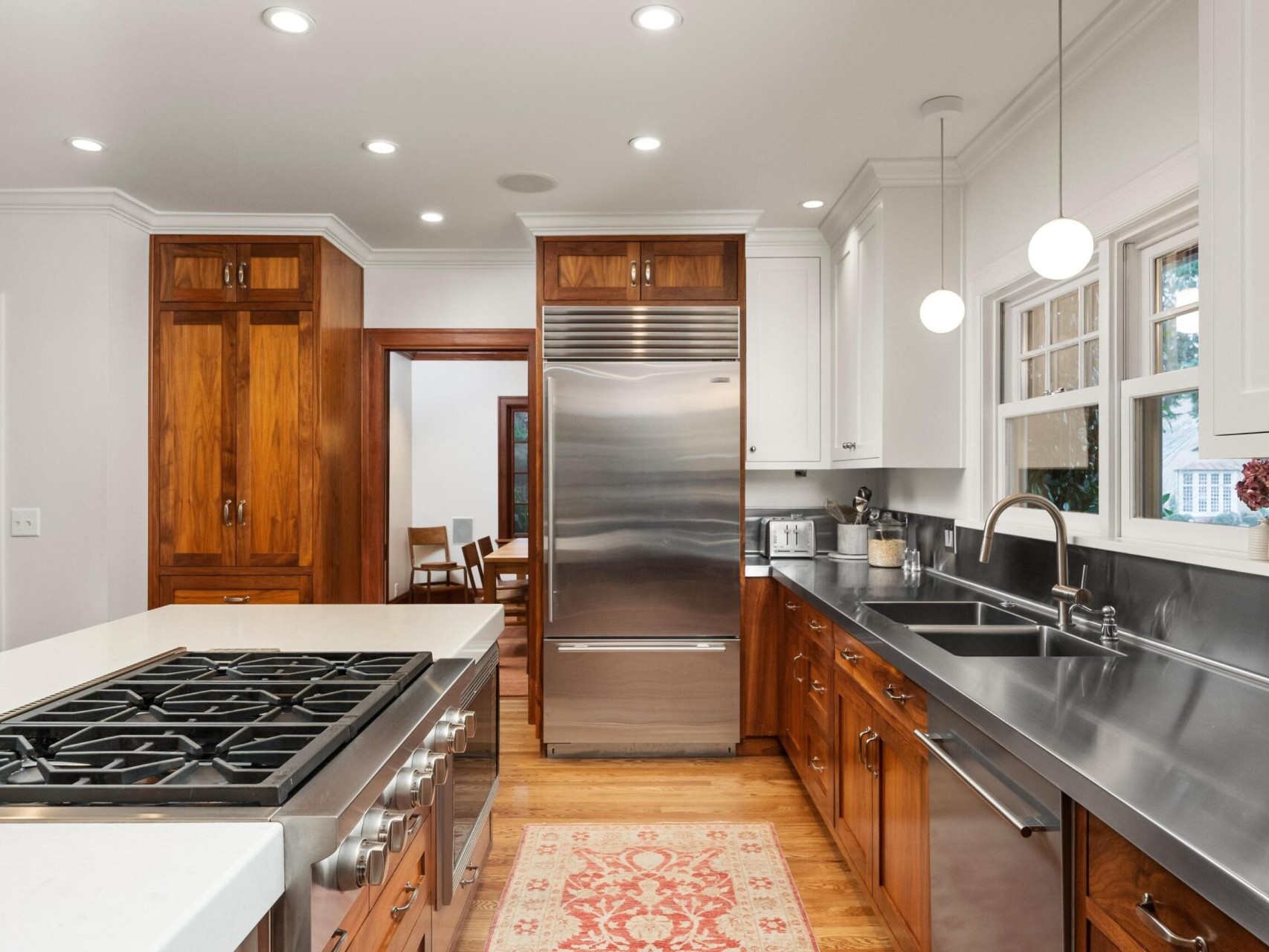 A modern kitchen featuring wooden cabinets, a stainless steel fridge and stove. A large island with a gas cooktop, double sink beneath two windows, and pendant lights overhead enhance its charm. This Portland Oregon Real Estate gem boasts a patterned rug on the wooden floor, making it truly inviting.