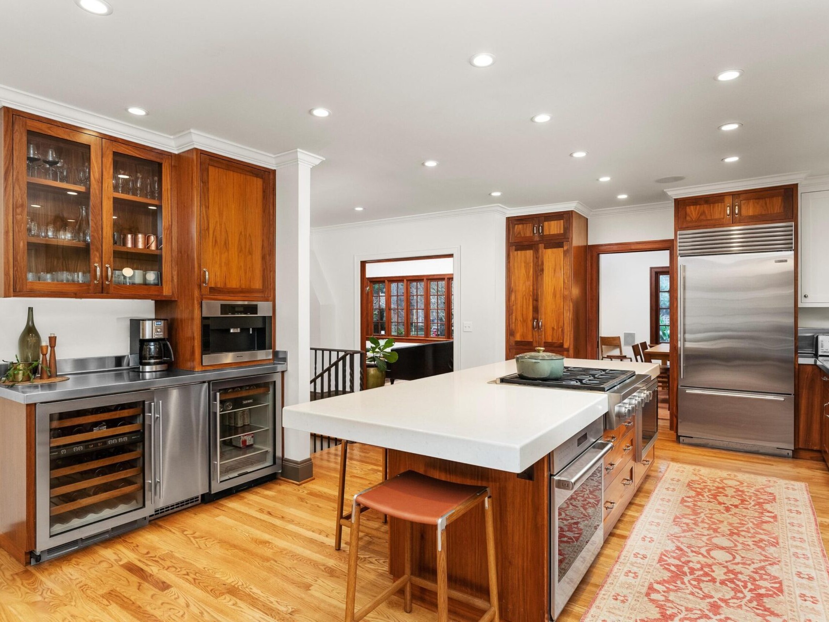 Spacious kitchen with wooden cabinetry, stainless steel appliances, and a center island with barstool on the hardwood floors. A decorative rug extends along the floor, while glass-front cabinets display glassware. This bright, well-lit space offers modern design, perfect for Portland Oregon real estate seekers.