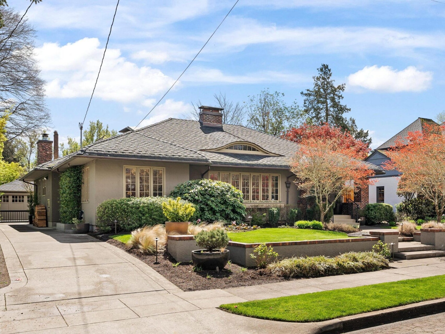 A charming suburban home in the Portland real estate market features a manicured front lawn and driveway. Its gray facade, large windows, and neatly landscaped garden with bushes and trees complement the clear blue sky, making it a gem for any top real estate agent to showcase.