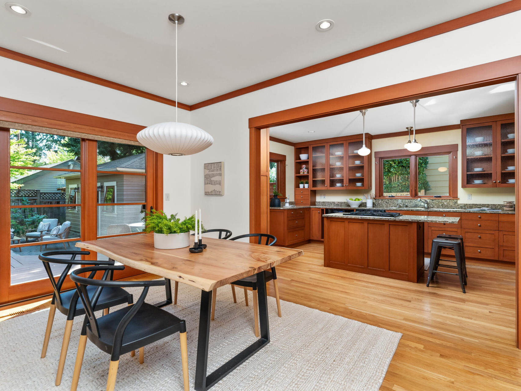 A bright, open dining room showcases a wooden table and black chairs on a light rug, perfect for entertaining. A large sliding glass door leads to the deck outside. The kitchen boasts wooden cabinets, granite countertops, and pendant lights—key highlights in this Portland Oregon real estate gem.