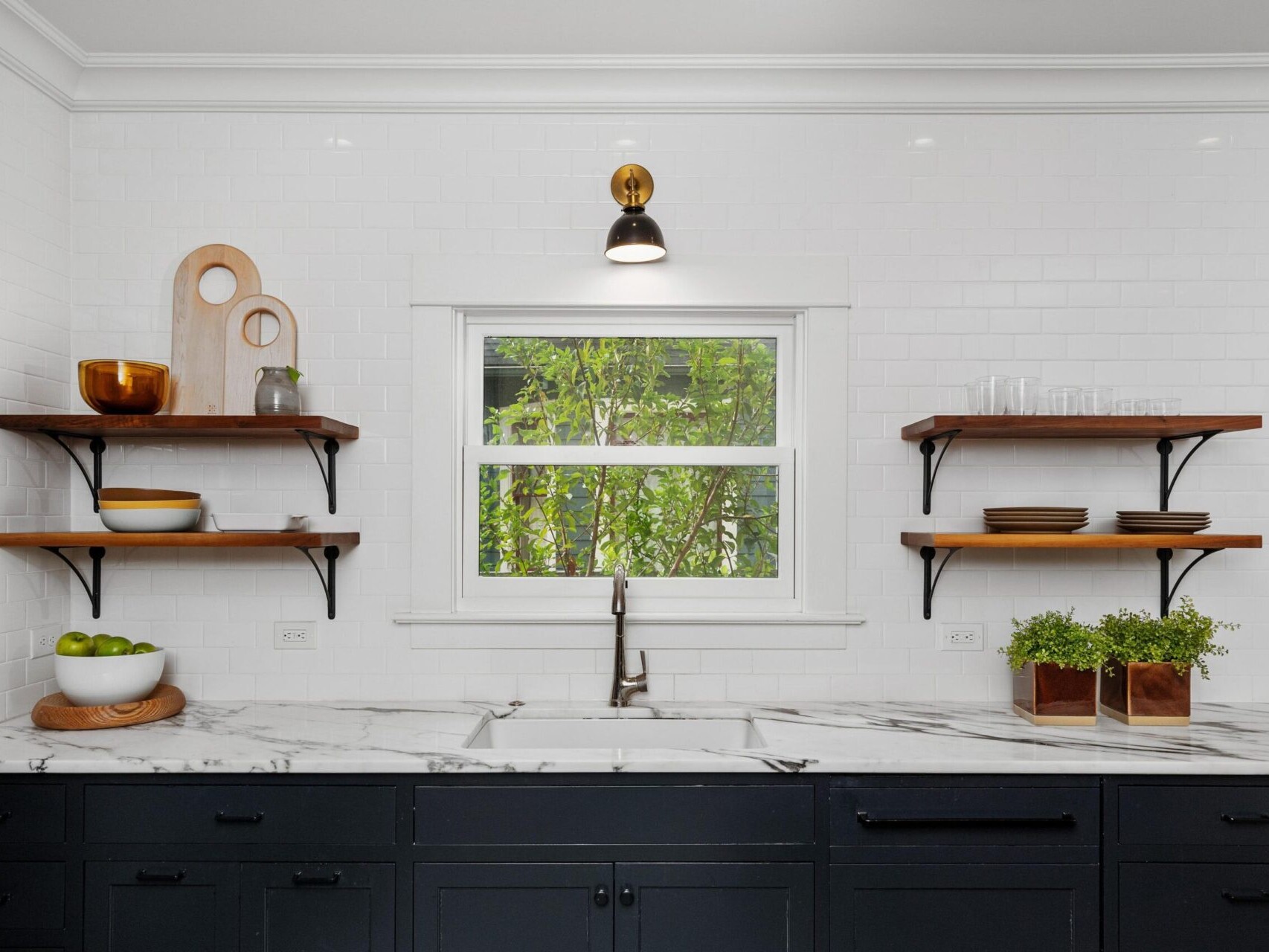 A modern kitchen in Portland, Oregon boasts a black and white color scheme with a marble countertop and dark cabinets. Wooden shelves on either side display dishes and decor items. A central window invites natural light, framed by a single wall light above.