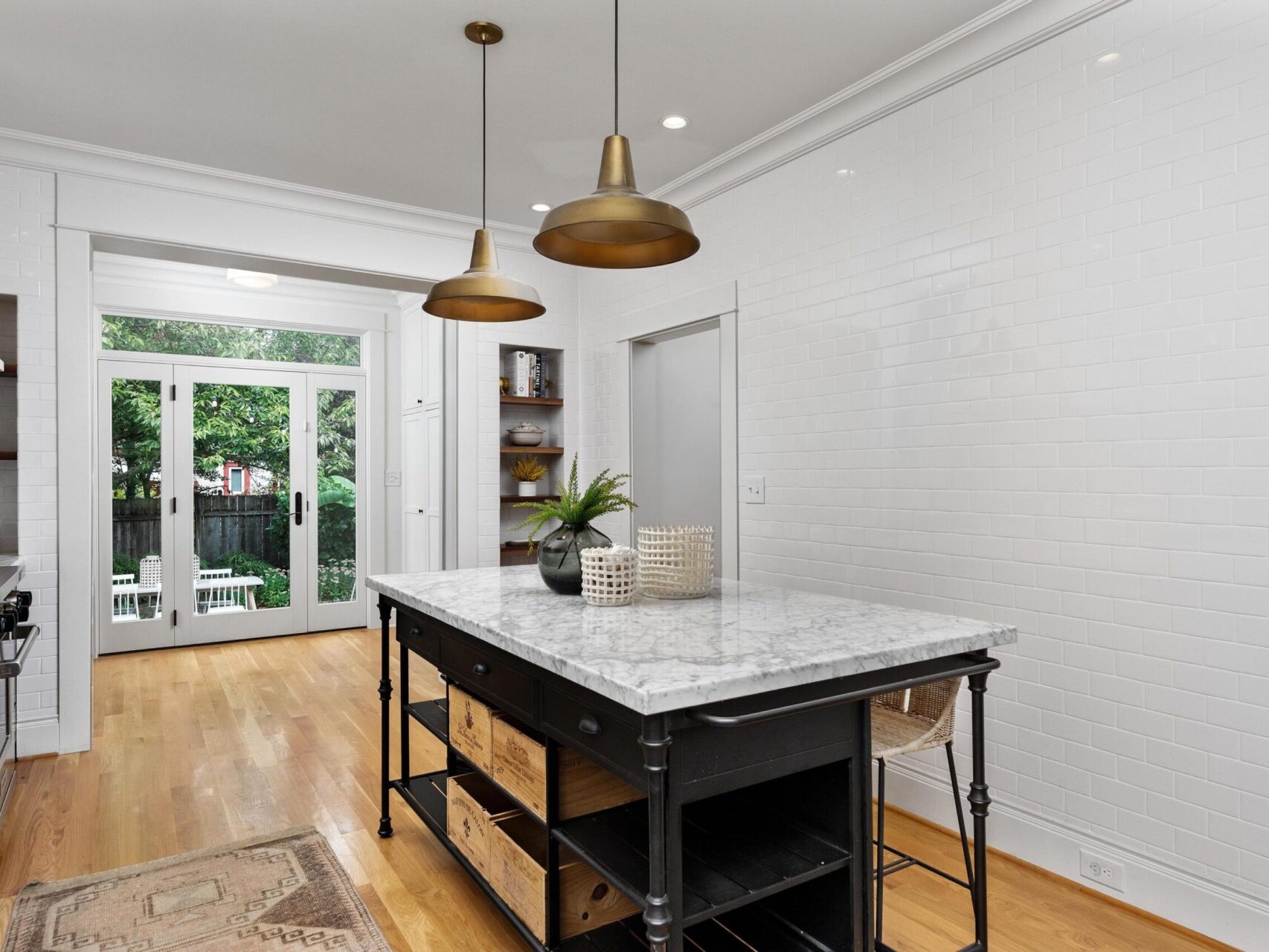 Modern kitchen with a marble-topped island featuring storage baskets and a potted plant. Pendant lights hang above. This Portland, Oregon space has white subway tile walls, hardwood floors, and French doors leading to a garden.