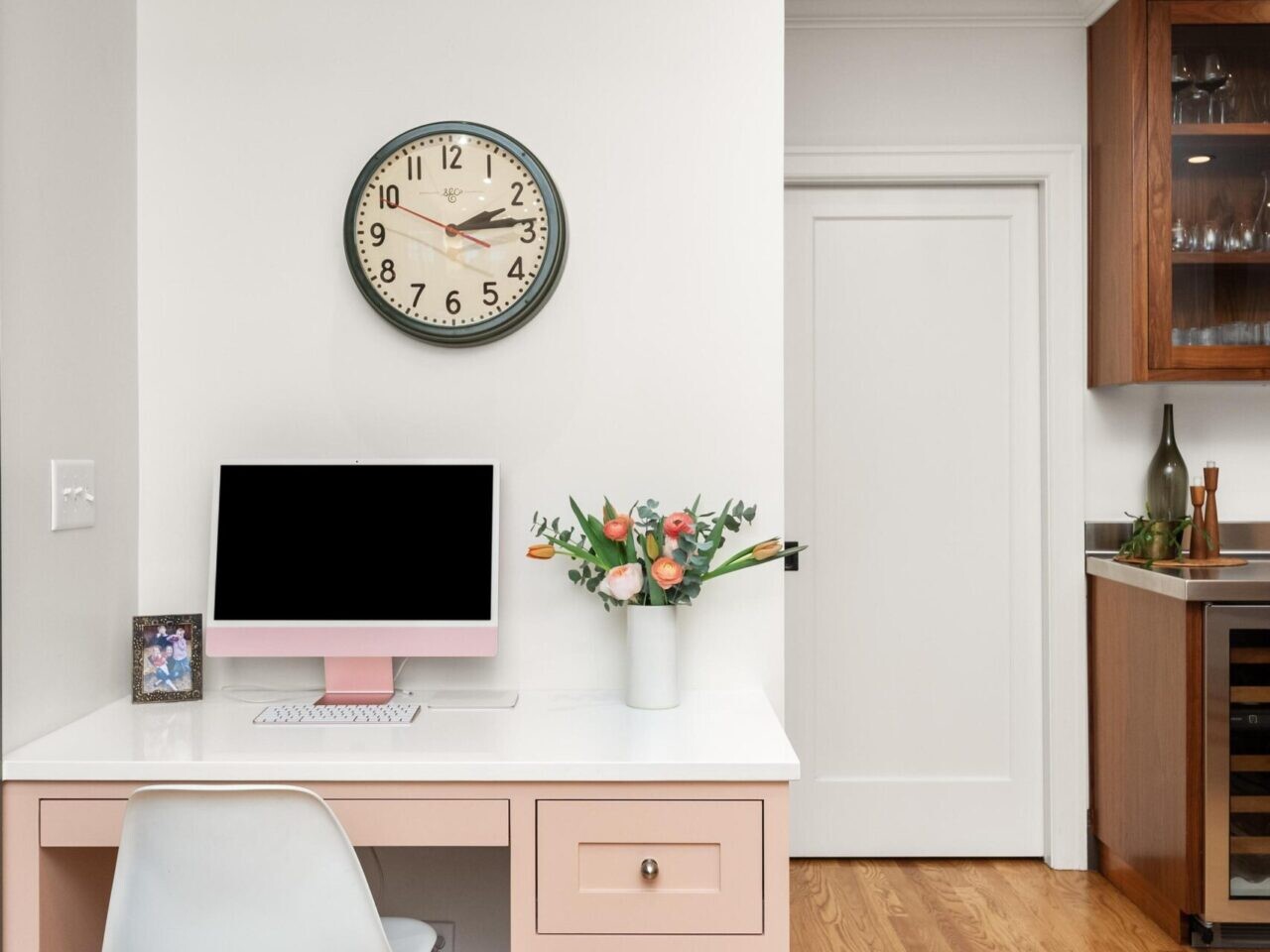 A home office setup perfect for a Portland Realtor features a pink desk with a computer, flowers in a vase, and a picture frame. A white chair is tucked in, and a wall clock hangs above. The space boasts hardwood flooring and stylish cabinetry, ideal for any Portland Oregon Real Estate professional.