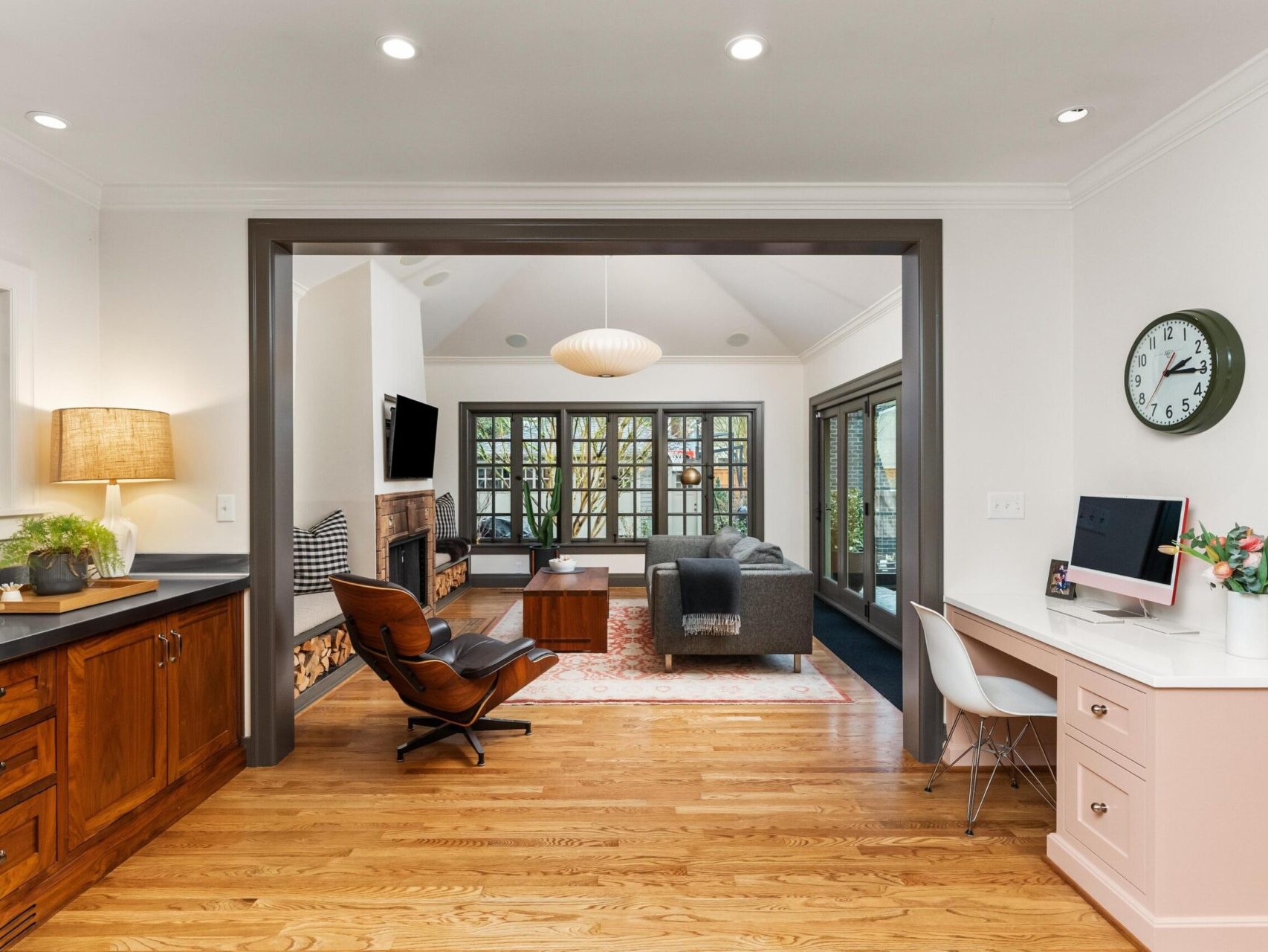 Spacious living area with wooden flooring, featuring a cozy seating arrangement with a gray sofa and armchair in front of a fireplace. A computer desk with a flower vase is on the right, and large windows let in natural light—a perfect find by your Portland Oregon real estate expert.