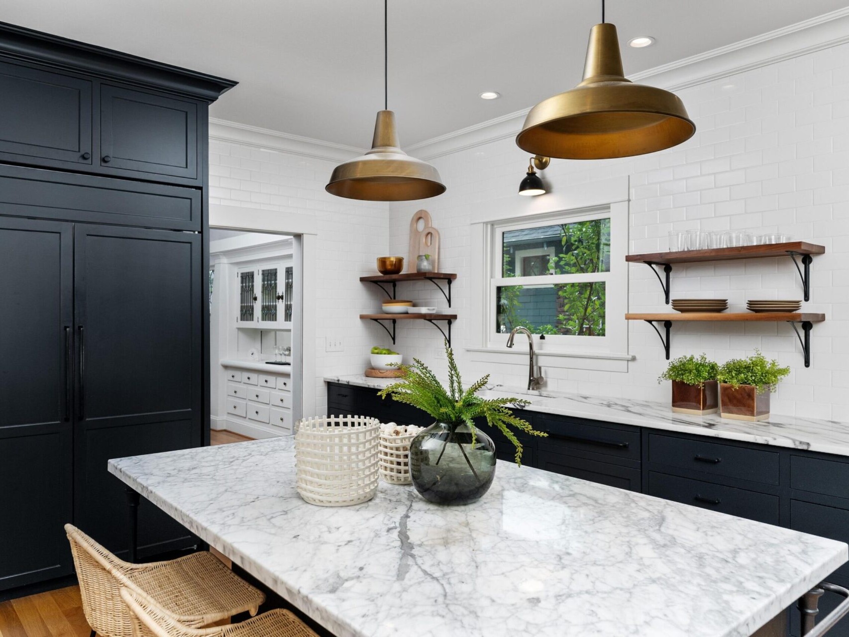 A modern kitchen in Portland, Oregon features white subway tile walls, black cabinets, and a marble island. Two bronze pendant lights hang above the island, adorned with plants and baskets. Wall shelves display dishes while a window floods the space with natural light.