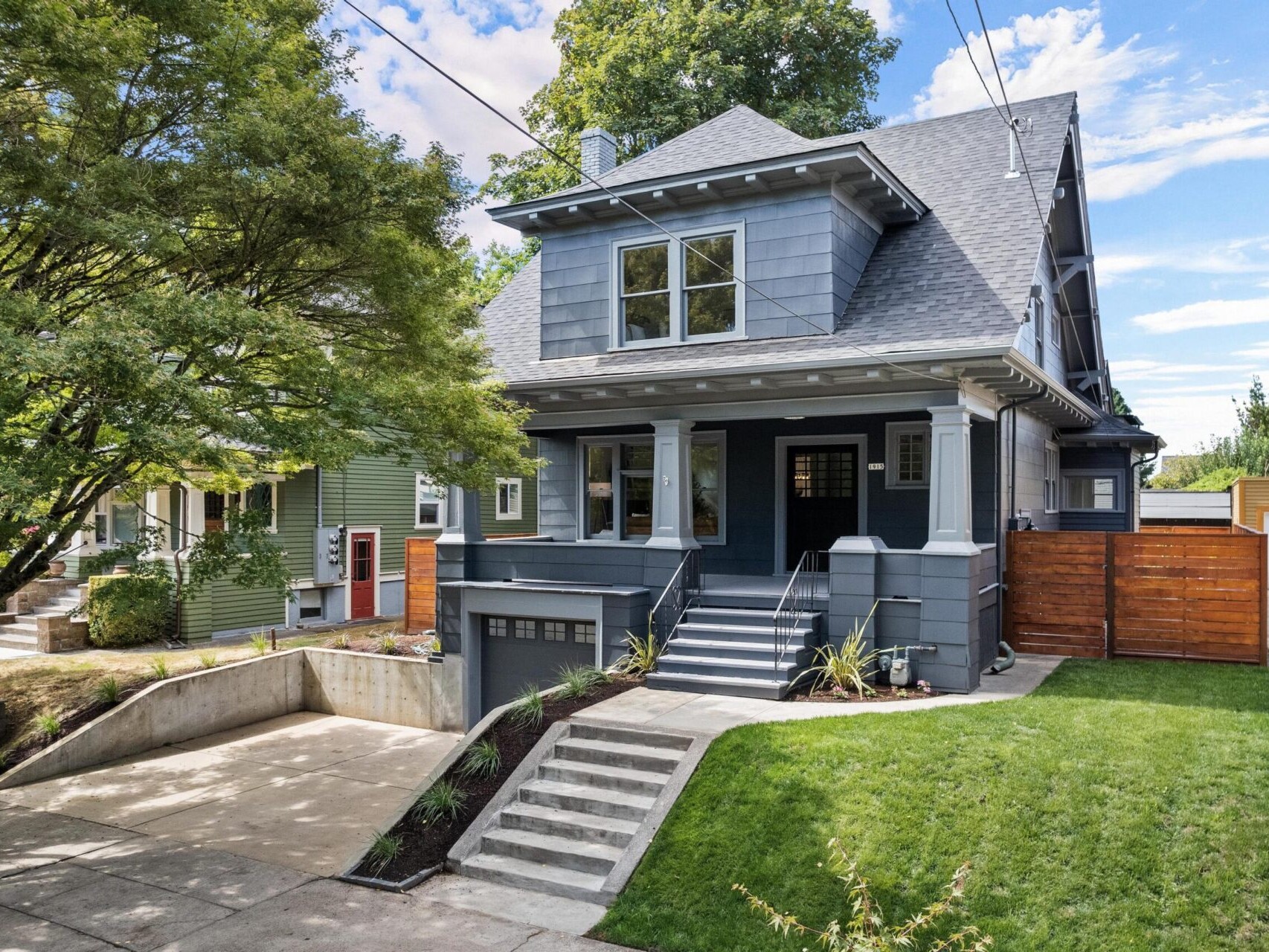 A two-story gray house with white trim stands on a quiet suburban street in Portland, Oregon. It features a steep roof, front porch, and landscaped yard with a driveway leading to a garage. Trees and a wooden fence frame the property under a partly cloudy sky.