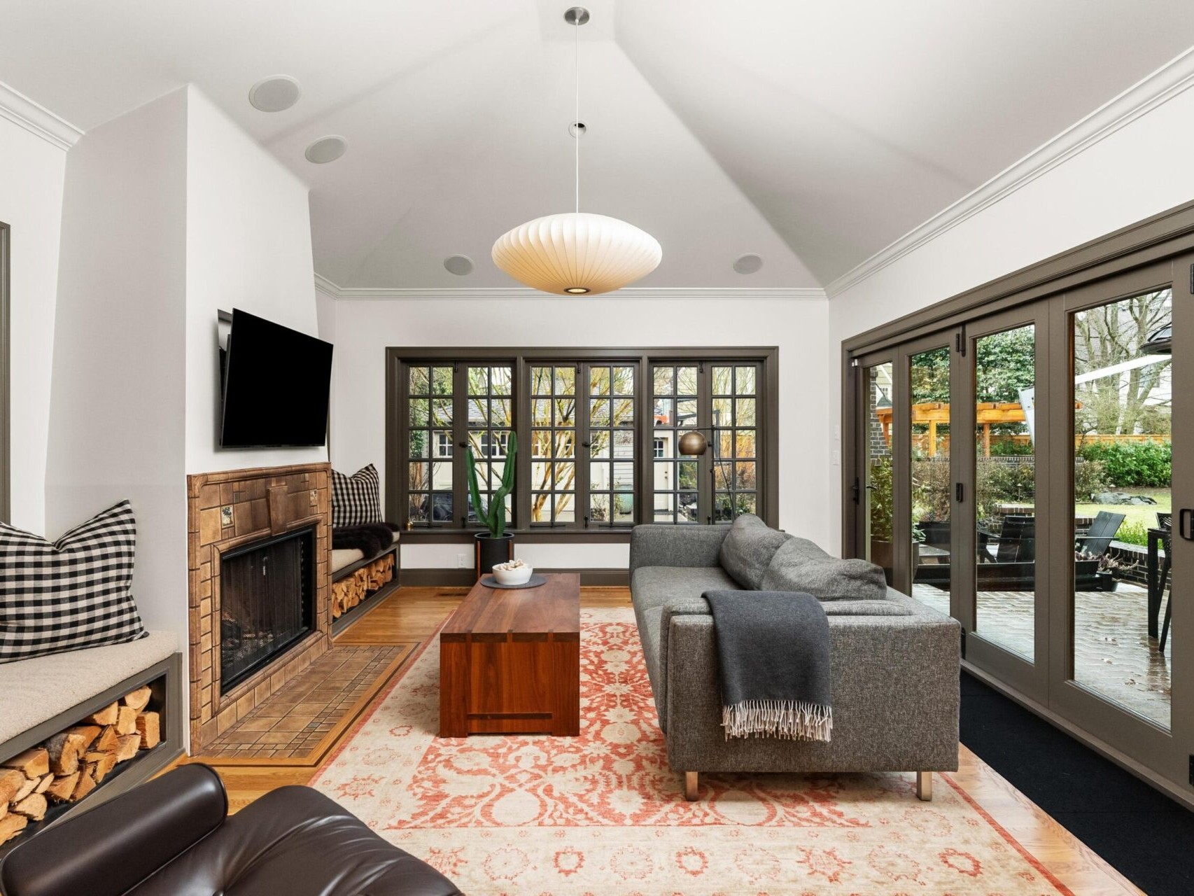 A cozy living room with a vaulted ceiling, featuring a gray sofa, armchair, wooden coffee table, and wall-mounted TV above the fireplace. Large windows and French doors open to an outdoor patio surrounded by greenery—a perfect gem in Portland Oregon real estate. An overhead light fixture brightens the space.