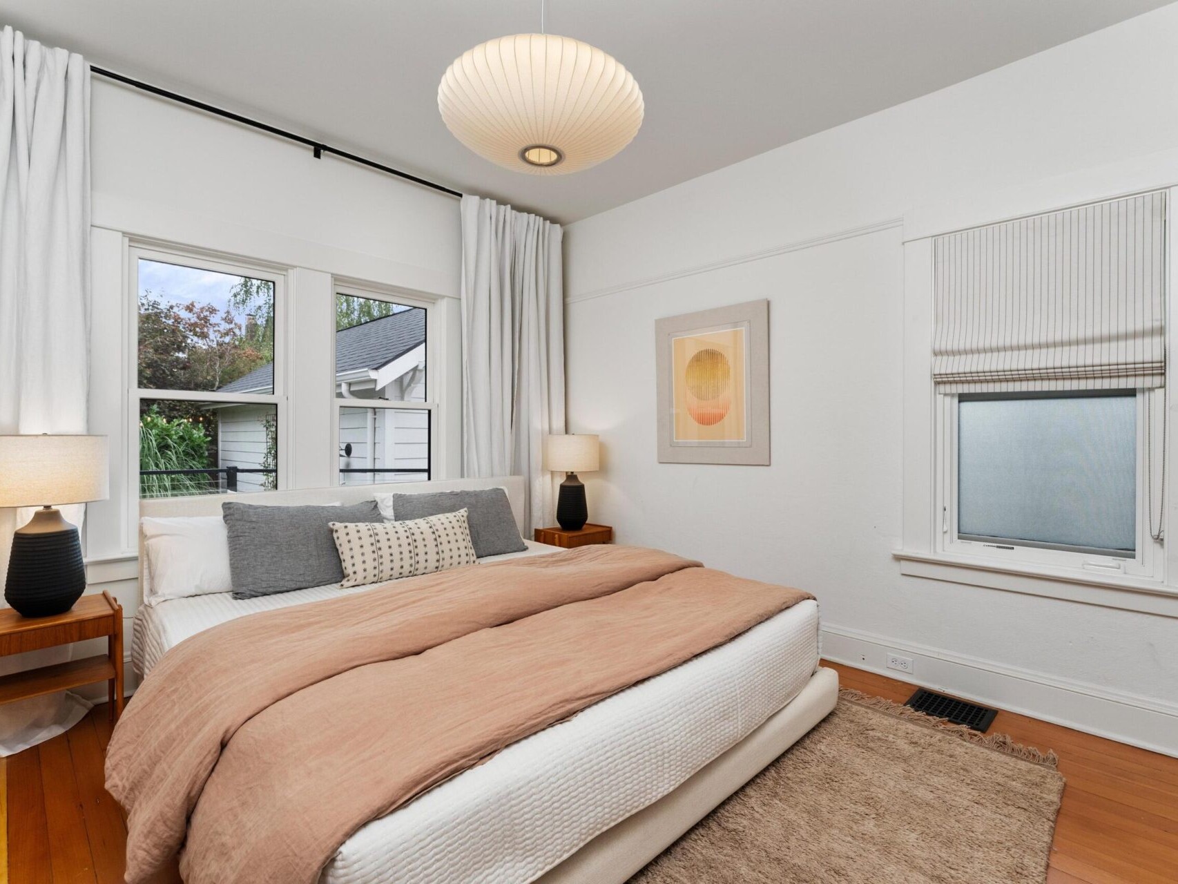 A cozy bedroom in Portland, Oregon, features a neatly made bed with a beige comforter and pillows. The room is lit by a modern pendant light and two lamps on wooden nightstands. A window with curtains and an abstract art piece adorn the walls, complemented by a small rug on the wooden floor.