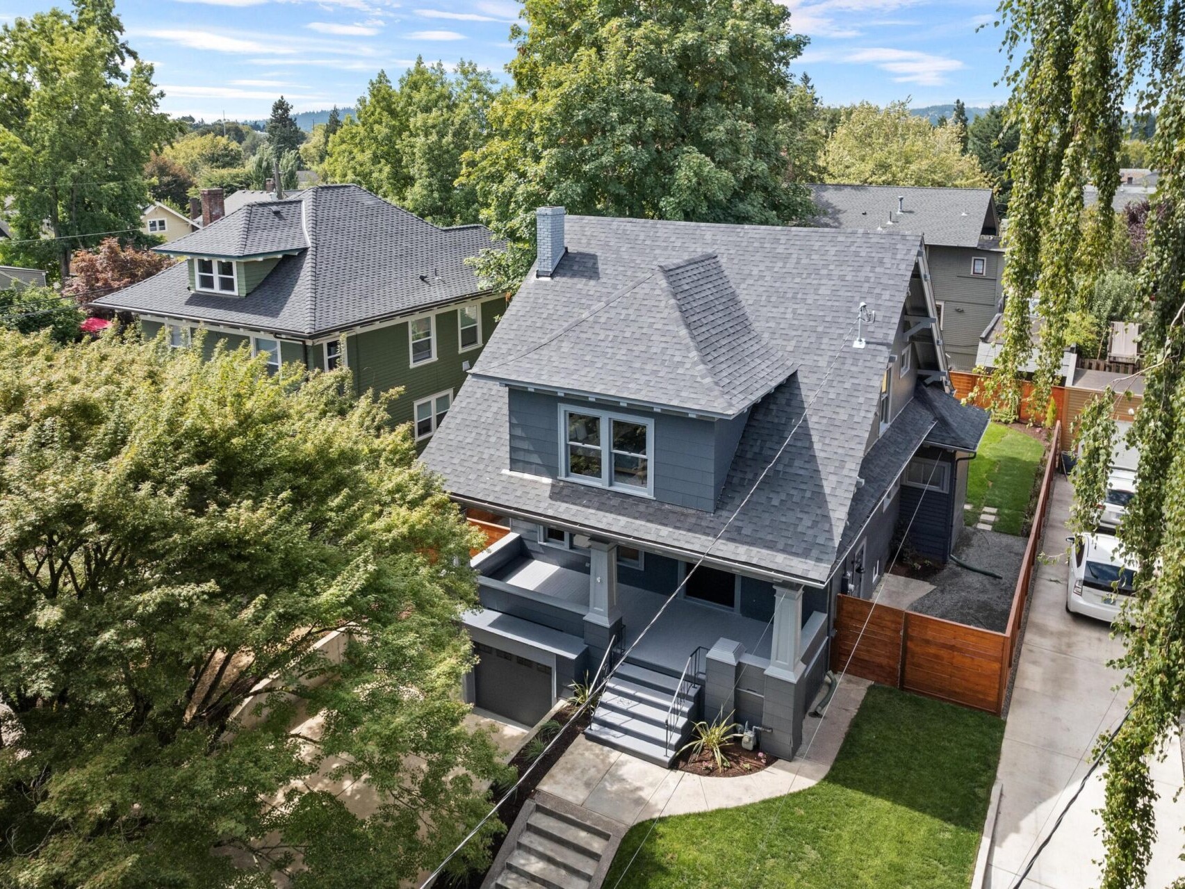Aerial view of a two-story gray house with a modern design in Portland, Oregon, surrounded by trees and greenery. A driveway and neatly trimmed lawn are visible, with neighboring homes in the background under a clear blue sky.