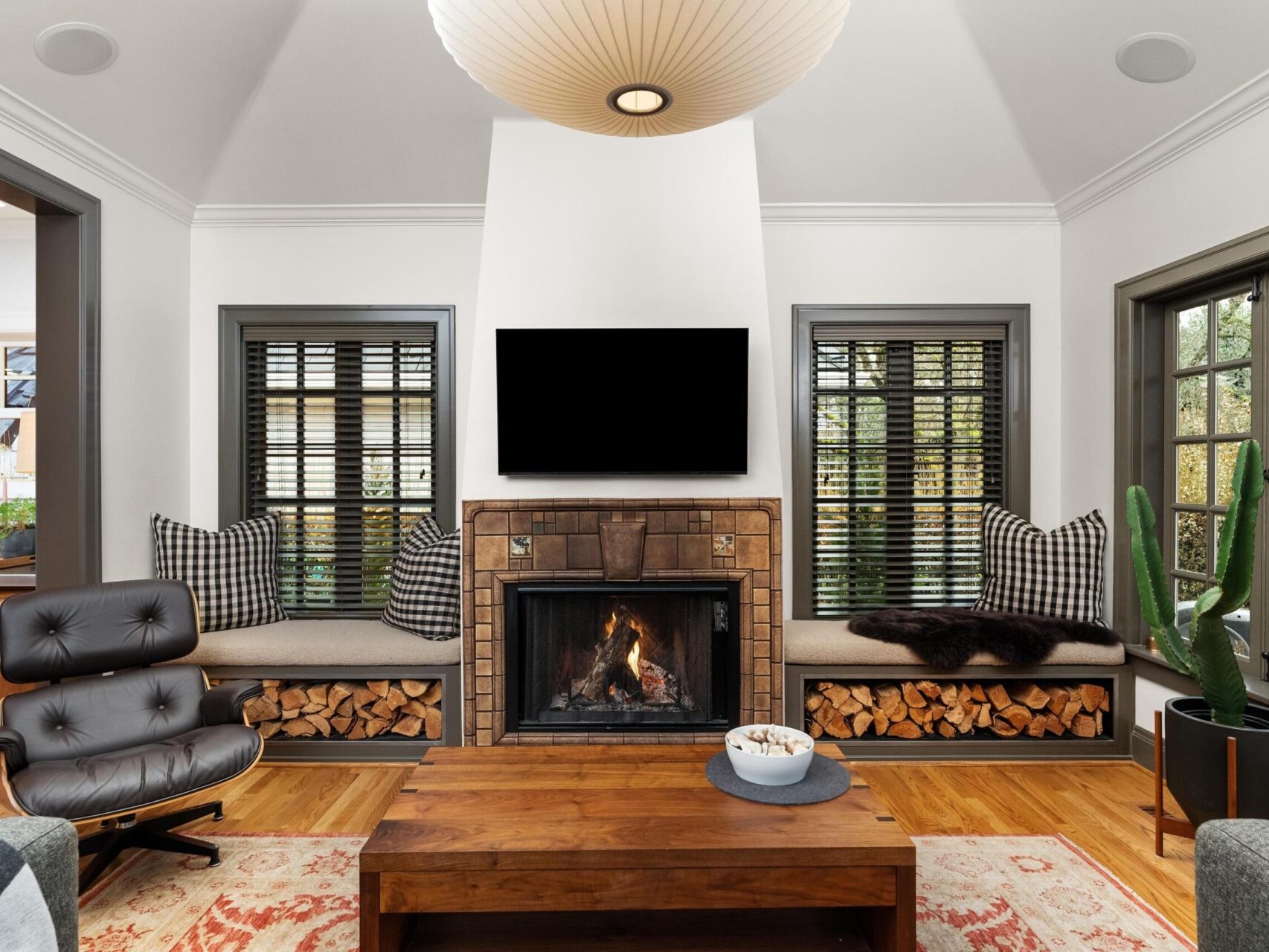 A cozy living room in a charming Portland, Oregon home features a fireplace with a wooden mantel, flanked by windows with bench seating. An Eames lounge chair sits on the left, and a wooden coffee table is centered on a patterned rug. A large ceiling light hangs above, perfect for any Portland Realtor to showcase.