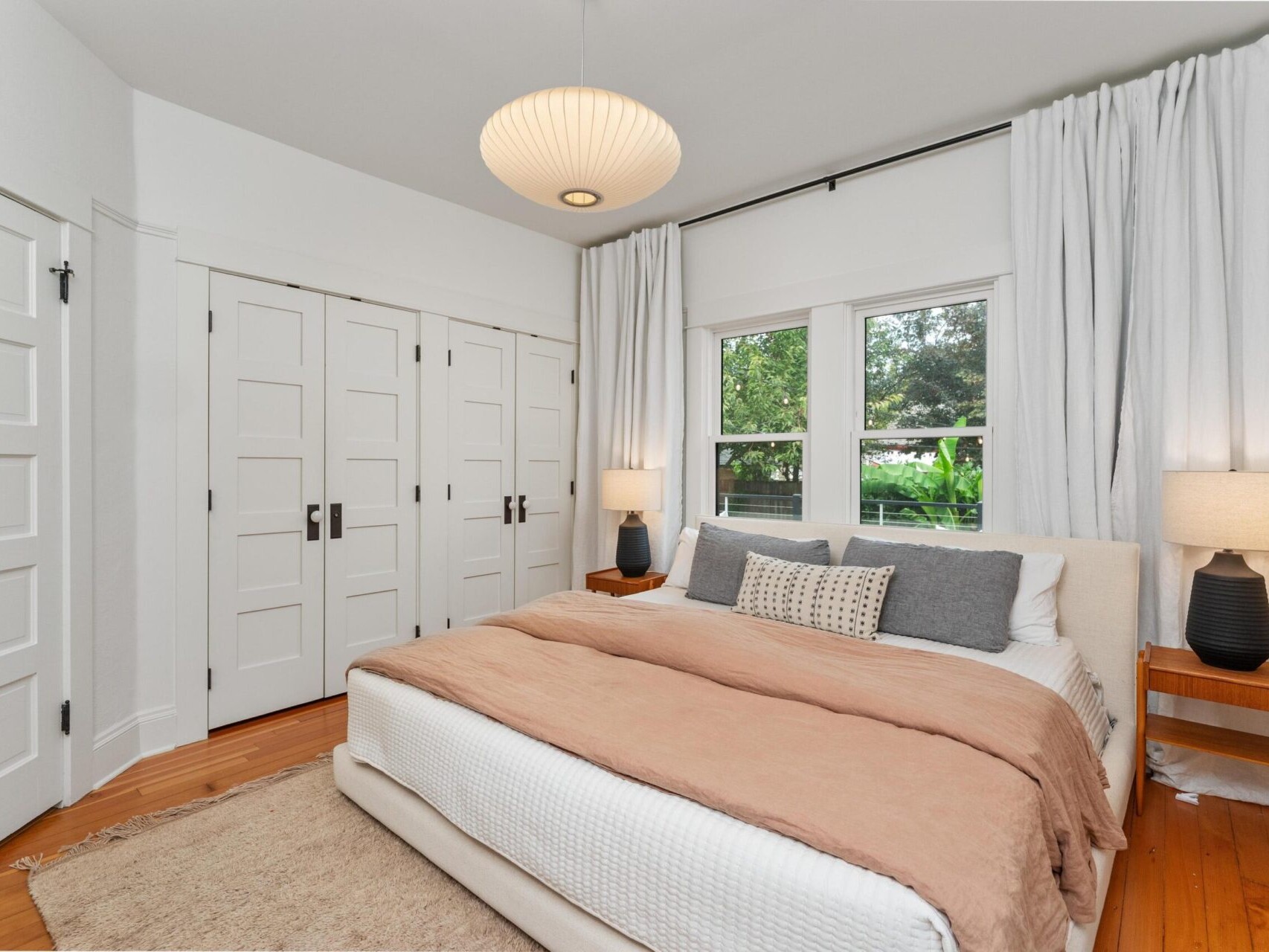 A cozy Portland, Oregon bedroom with a large bed featuring a beige comforter and decorative pillows. Two wooden nightstands with lamps flank the bed. The room boasts white walls, a light fixture above, and a window with white drapes revealing lush Pacific Northwest greenery outside.