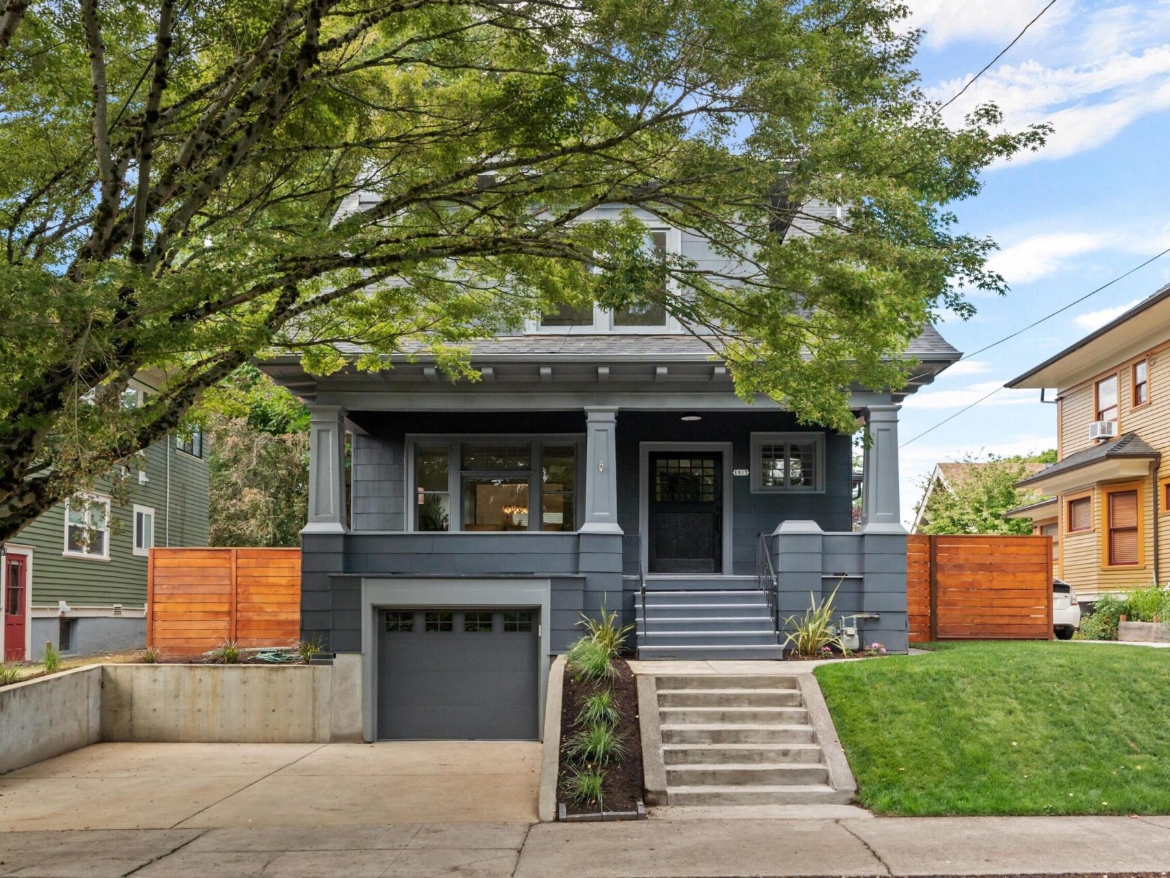 A charming two-story gray house in Portland, Oregon, boasts a covered porch surrounded by lush greenery and trees. It features a garage, a concrete driveway, and a neatly manicured lawn. A wooden fence partially encloses the yard under the clear blue sky.
