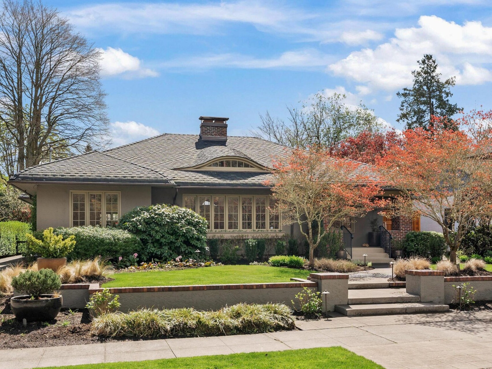 A charming craftsman-style house with a gray roof and beige walls is surrounded by neatly landscaped gardens. Brightly colored trees and lush greenery frame the scene under a partly cloudy blue sky, an ideal find in Portland Oregon real estate.