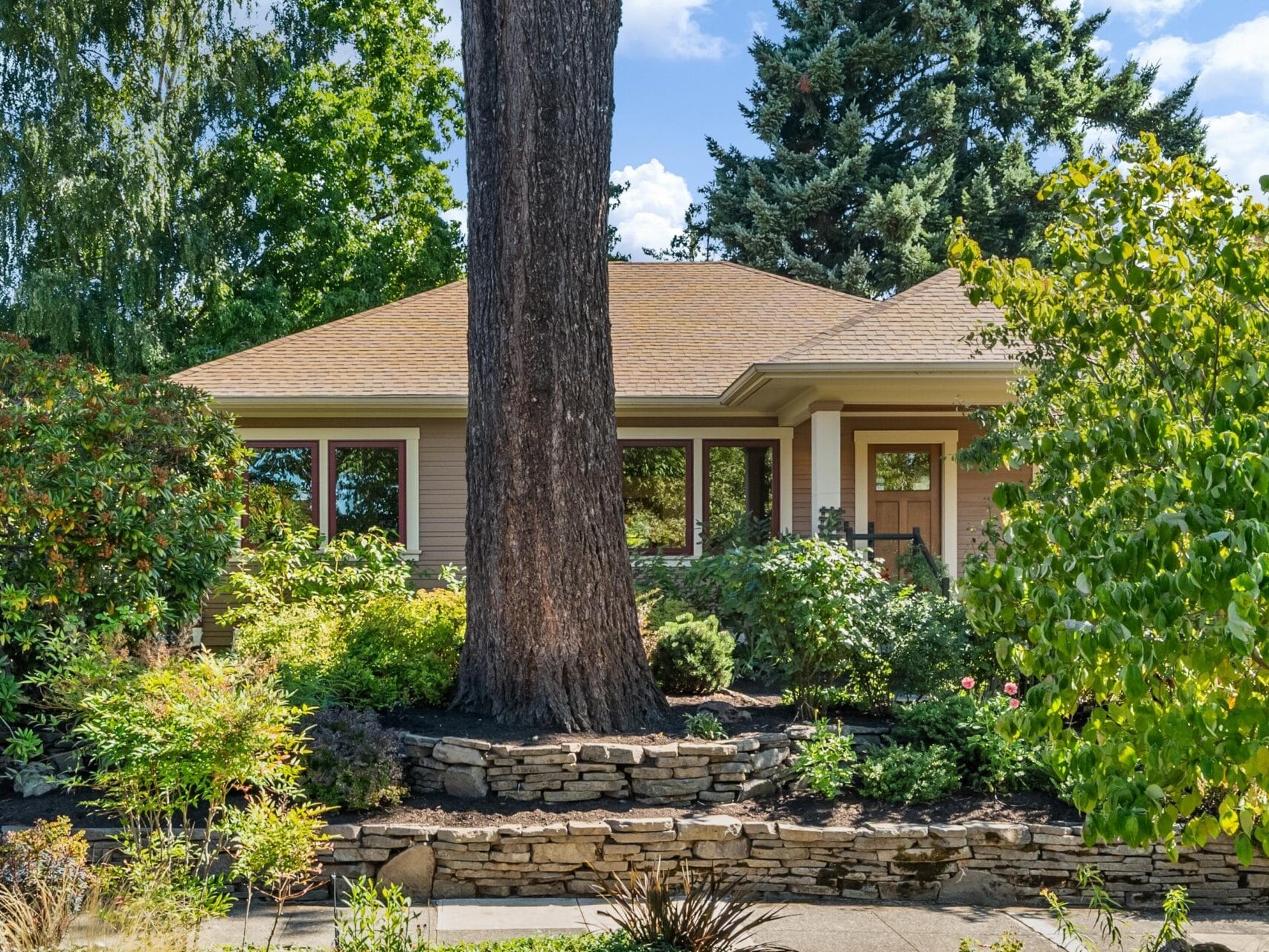A charming single-story house with a beige exterior is nestled in lush Portland, Oregon greenery. A large tree stands prominently in front, while a stone wall lines the landscaped garden. Under the clear blue sky, it embodies serene living in this picturesque locale.