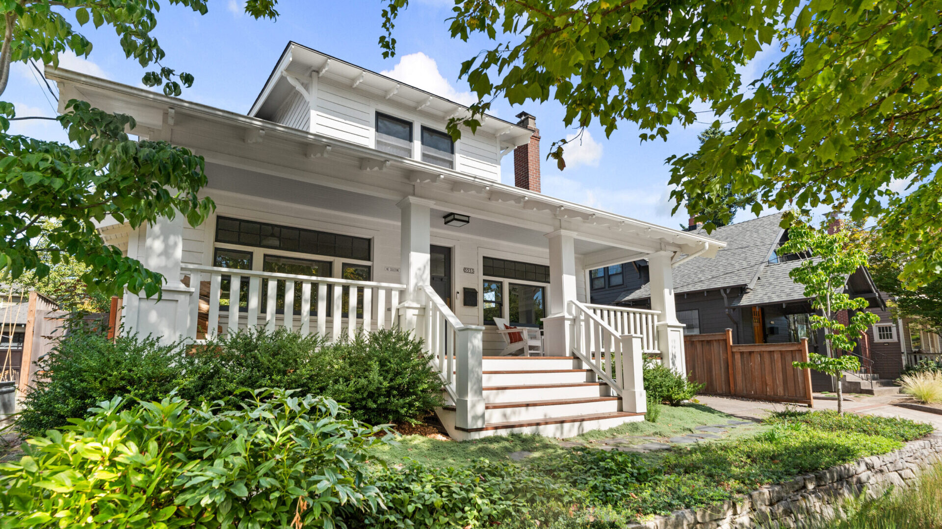 A charming white two-story house with a spacious porch surrounded by lush greenery and trees offers the quintessential Portland, Oregon, real estate charm. The blue sky and sunlight create a welcoming atmosphere, with steps leading up to the porch accented by white railings and cozy seating.