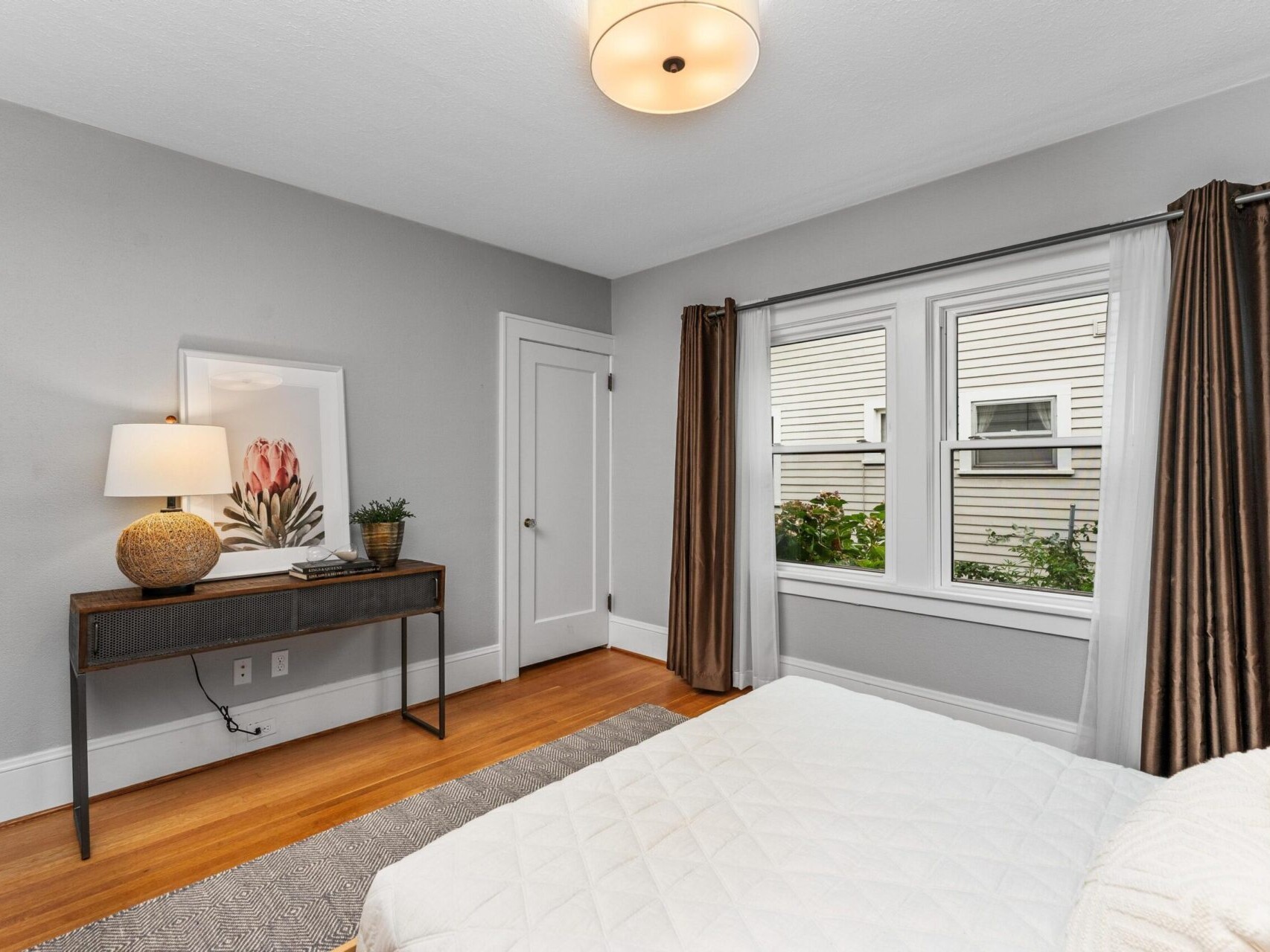 A cozy bedroom with light gray walls, hardwood floors, and a white bedspread. A wooden table holding a lamp and framed art is visible. Two windows with brown curtains allow natural light in. A small rug is on the floor beside the bed.