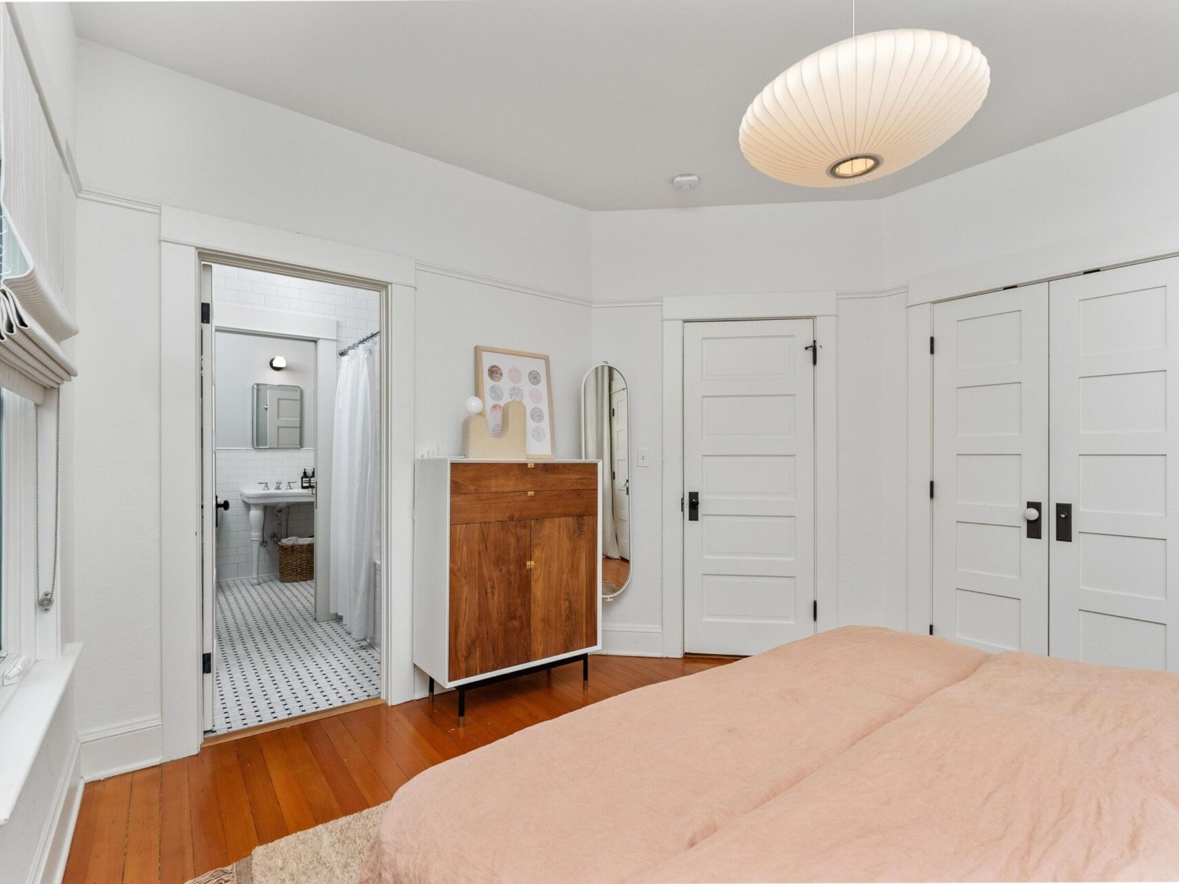 A cozy Portland, Oregon bedroom features white walls and a wooden floor. A bed with a peach duvet sits in the foreground alongside a wooden dresser, round mirror, and a view into a bathroom with black and white tile. A pendant lamp adds charm from the ceiling.