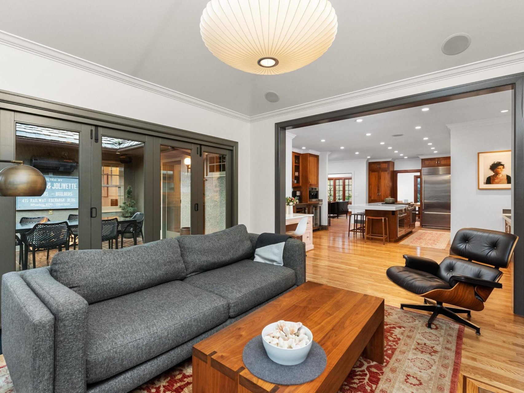 A modern living room features a gray sofa, wooden coffee table, and black lounge chair. Glass doors open to a patio with outdoor seating. The space connects to a kitchen with wooden cabinets and an island. A large pendant light hangs from the ceiling—a stunning find in Portland Oregon Real Estate.
