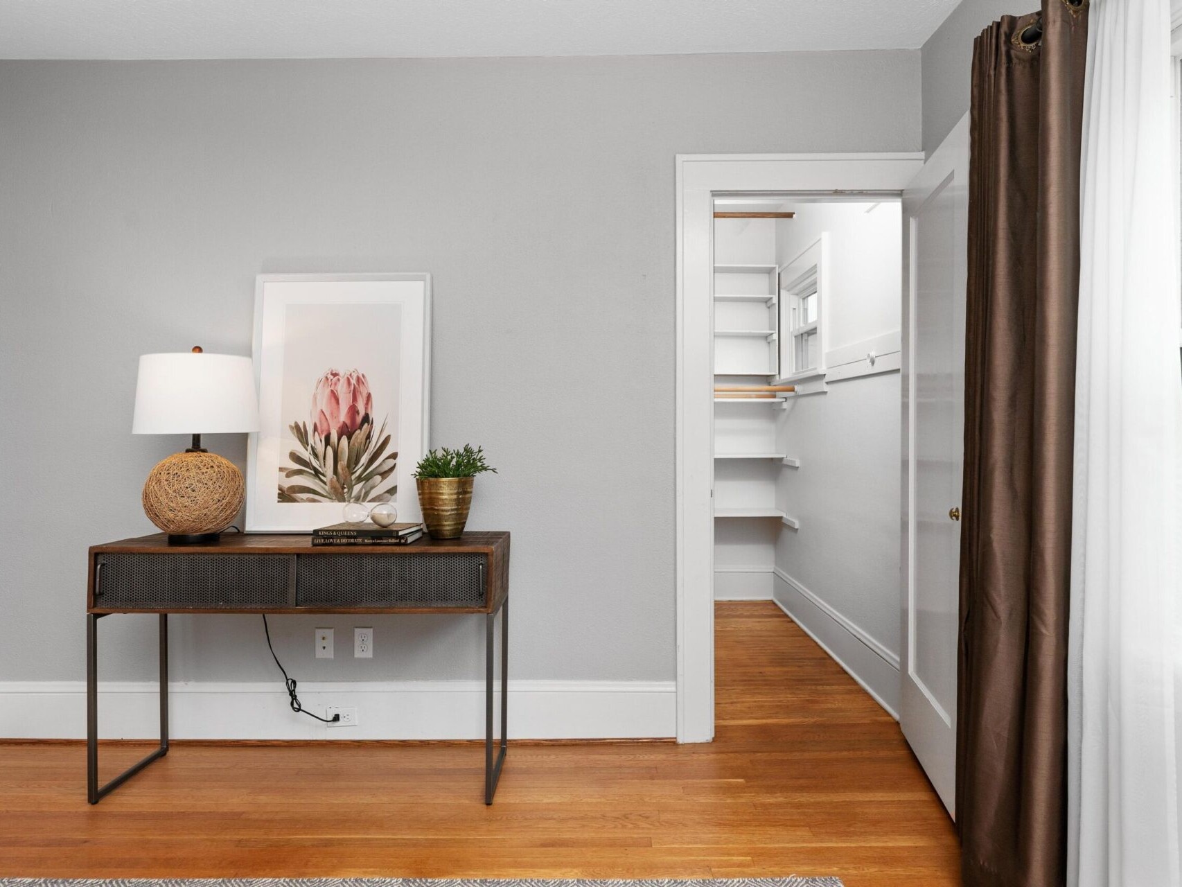 A minimalistic room with gray walls featuring a console table with a lamp, a decorative object, and a framed floral artwork. It overlooks a small, organized walk-in closet with shelves, and has hardwood floors and a large window with brown curtains.