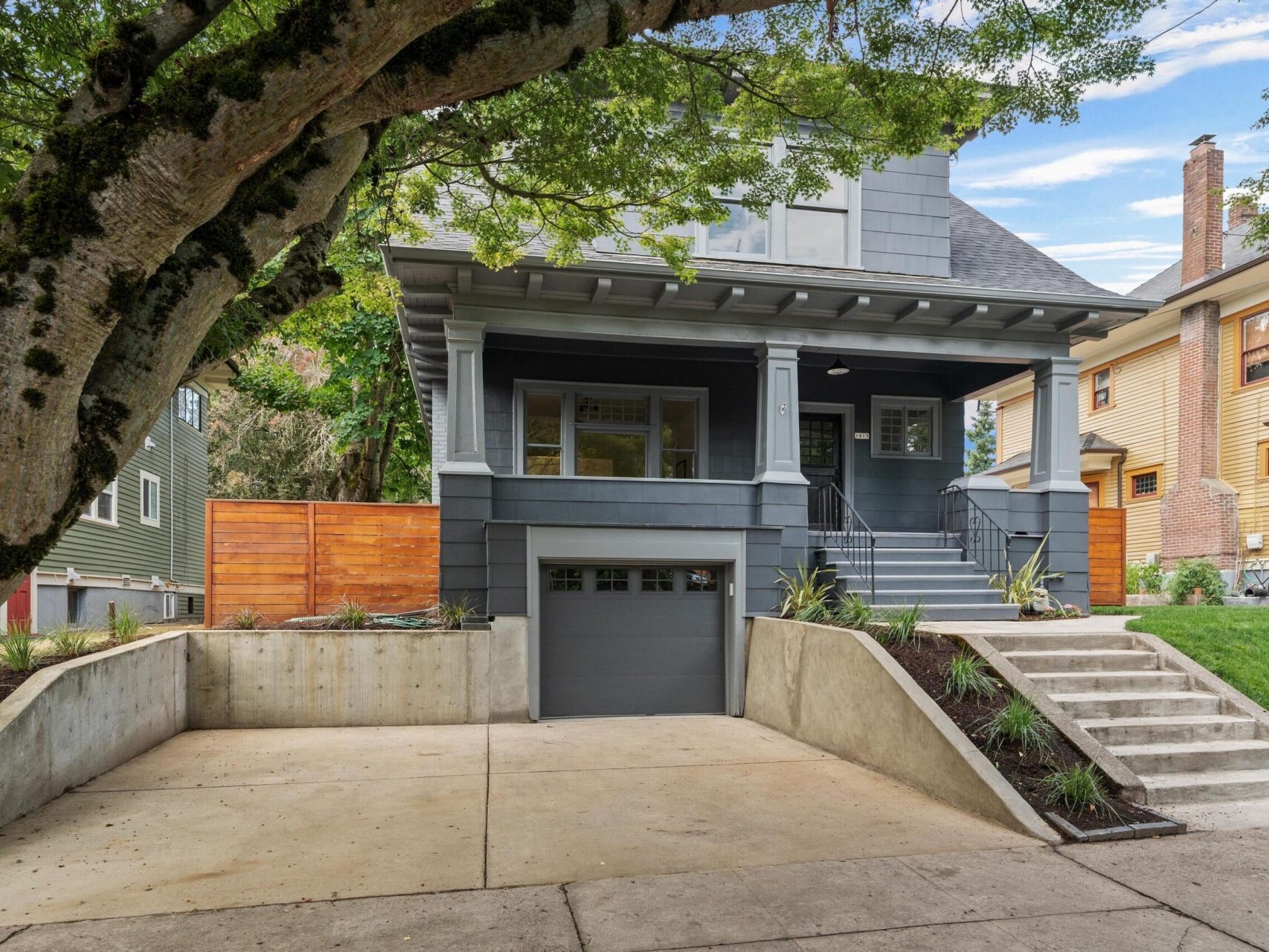 A modern gray two-story house in Portland, Oregon, features a spacious front porch and steps leading to the entrance. It boasts a large tree in the foreground and a driveway leading to a garage, surrounded by lush landscaping of grass and plants.
