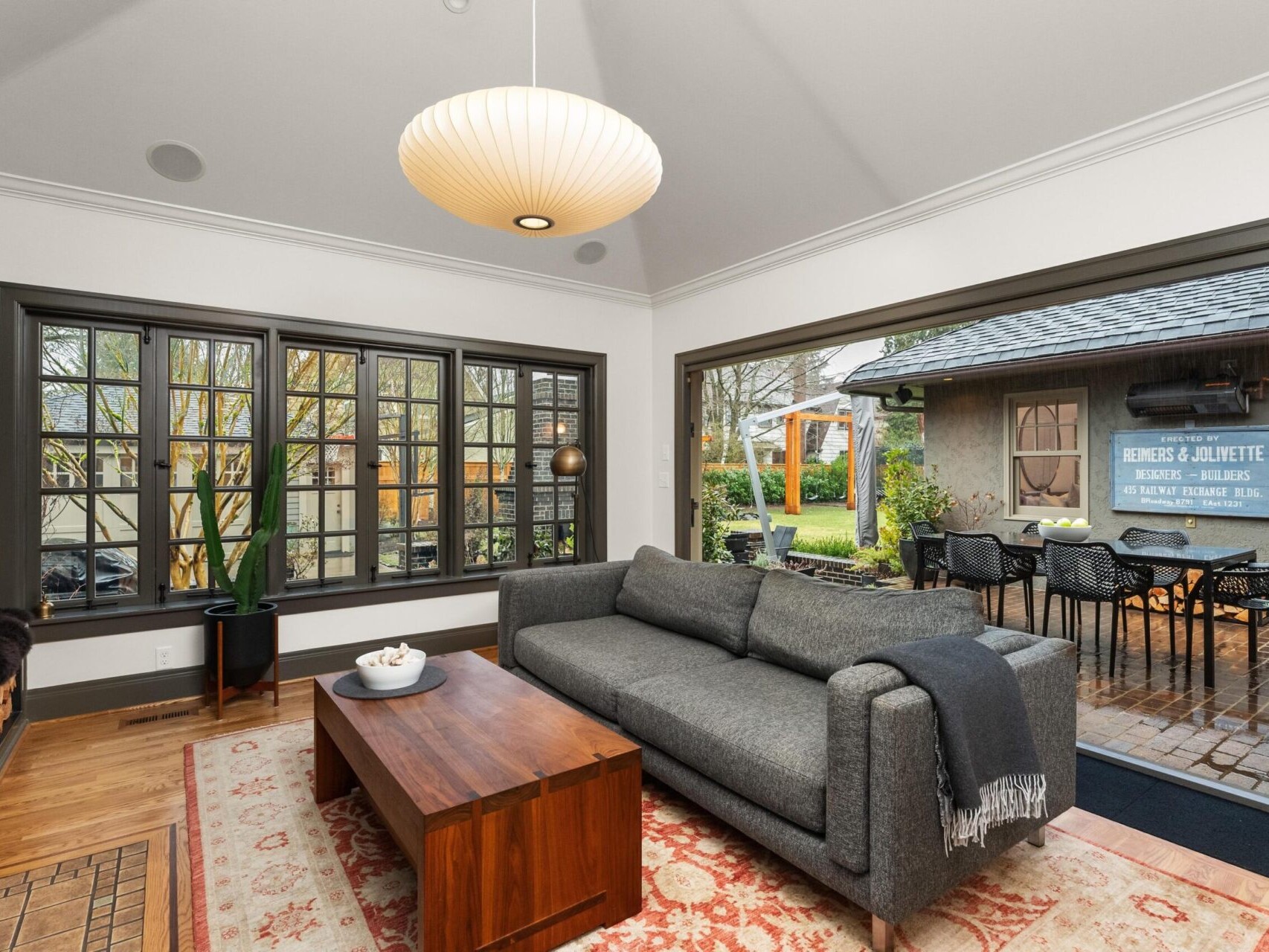 A cozy living room featuring a gray sofa, wooden coffee table, and a large area rug. The space boasts a wall-mounted TV and large windows, offering views of the lush garden outside. Glass doors lead to a patio with dining set, embodying the charm of Portland Oregon real estate.