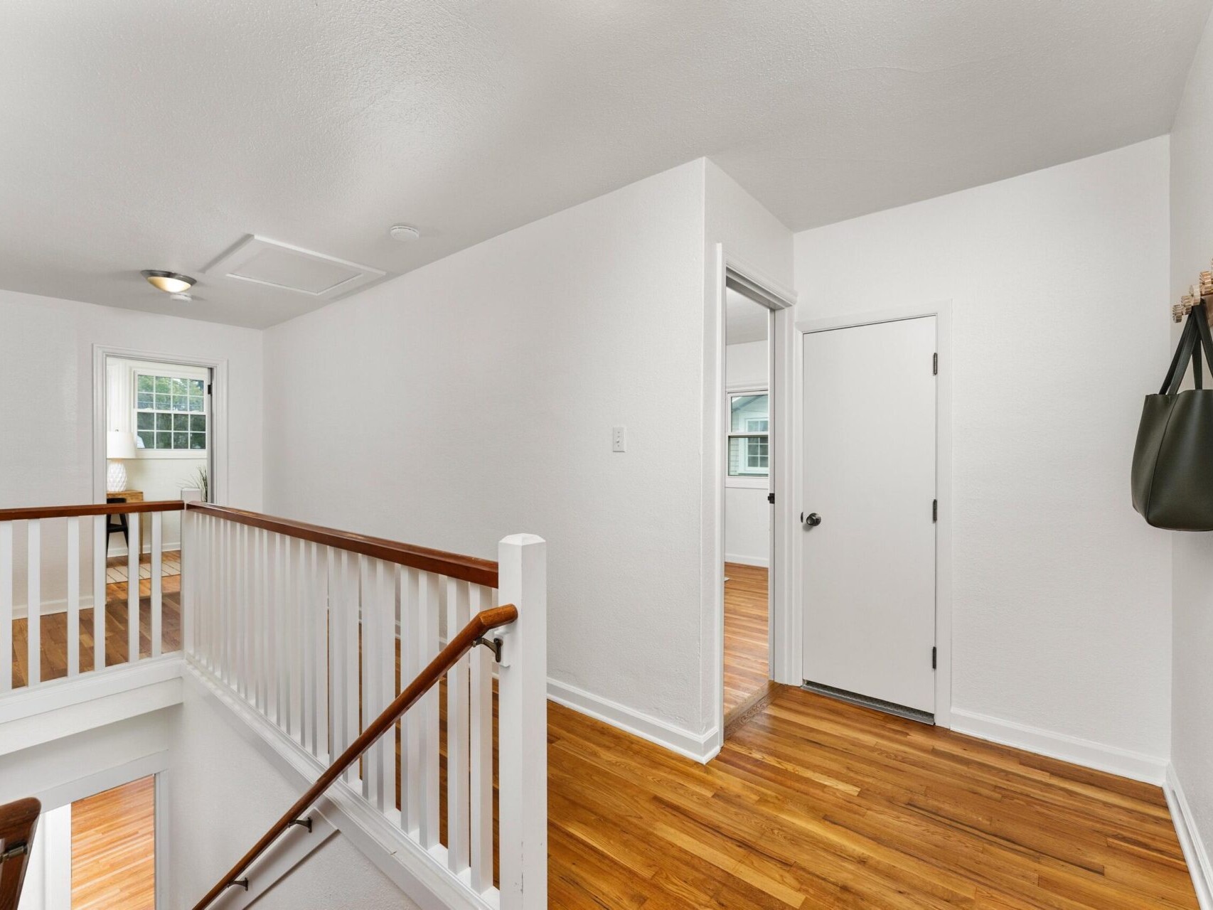 A bright hallway with white walls and wooden flooring. A staircase with white railings leads down. A green bag hangs on a wall hook to the right. A door and a room with natural light are visible in the background.