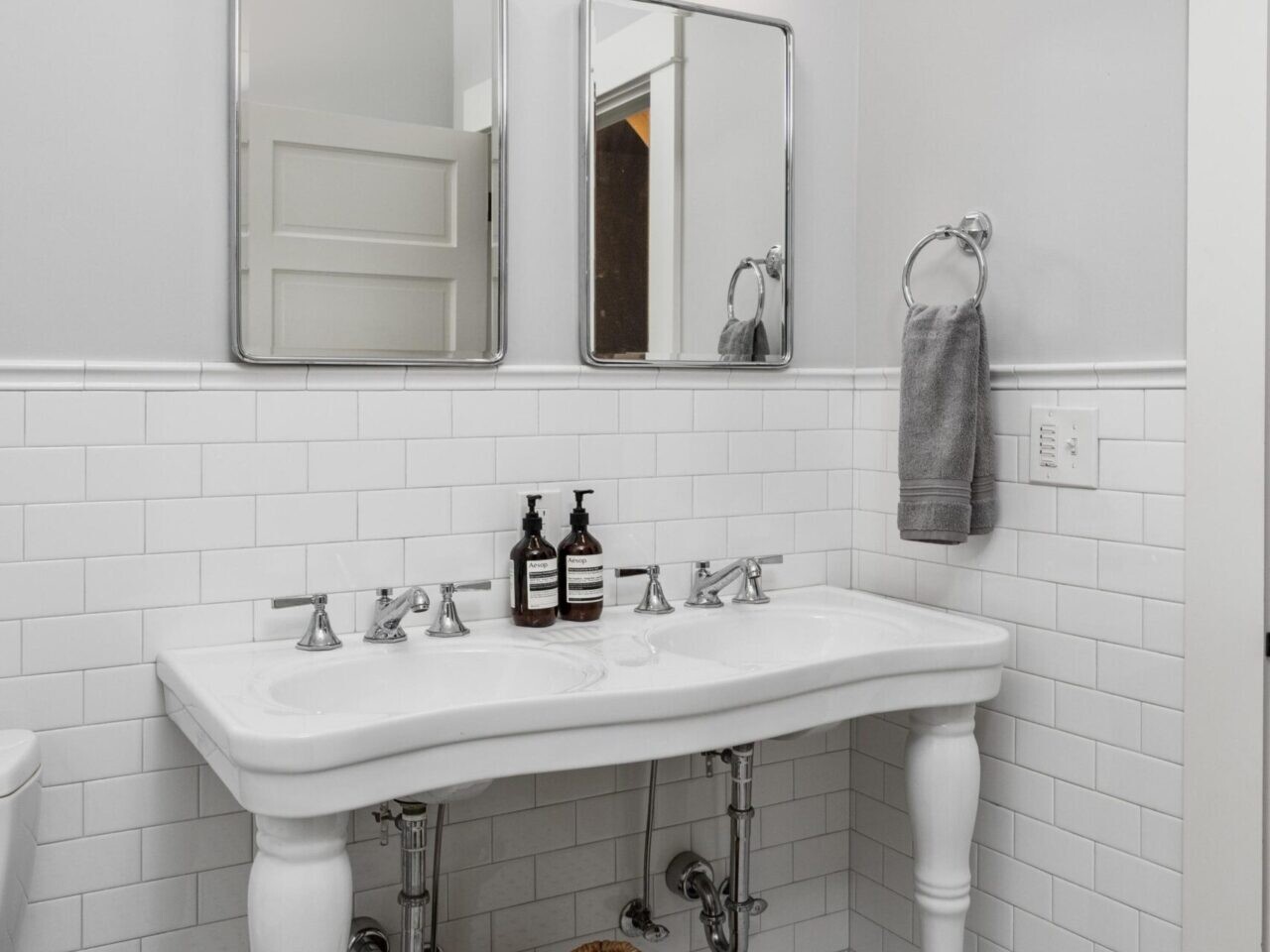 A Portland, Oregon-inspired bathroom features a classic white sink with dual faucets, two rectangular mirrors above, and two wall-mounted light fixtures. A wicker basket holds towels on the patterned tile floor, while a gray towel hangs on a hook, capturing the city's unique blend of style and simplicity.