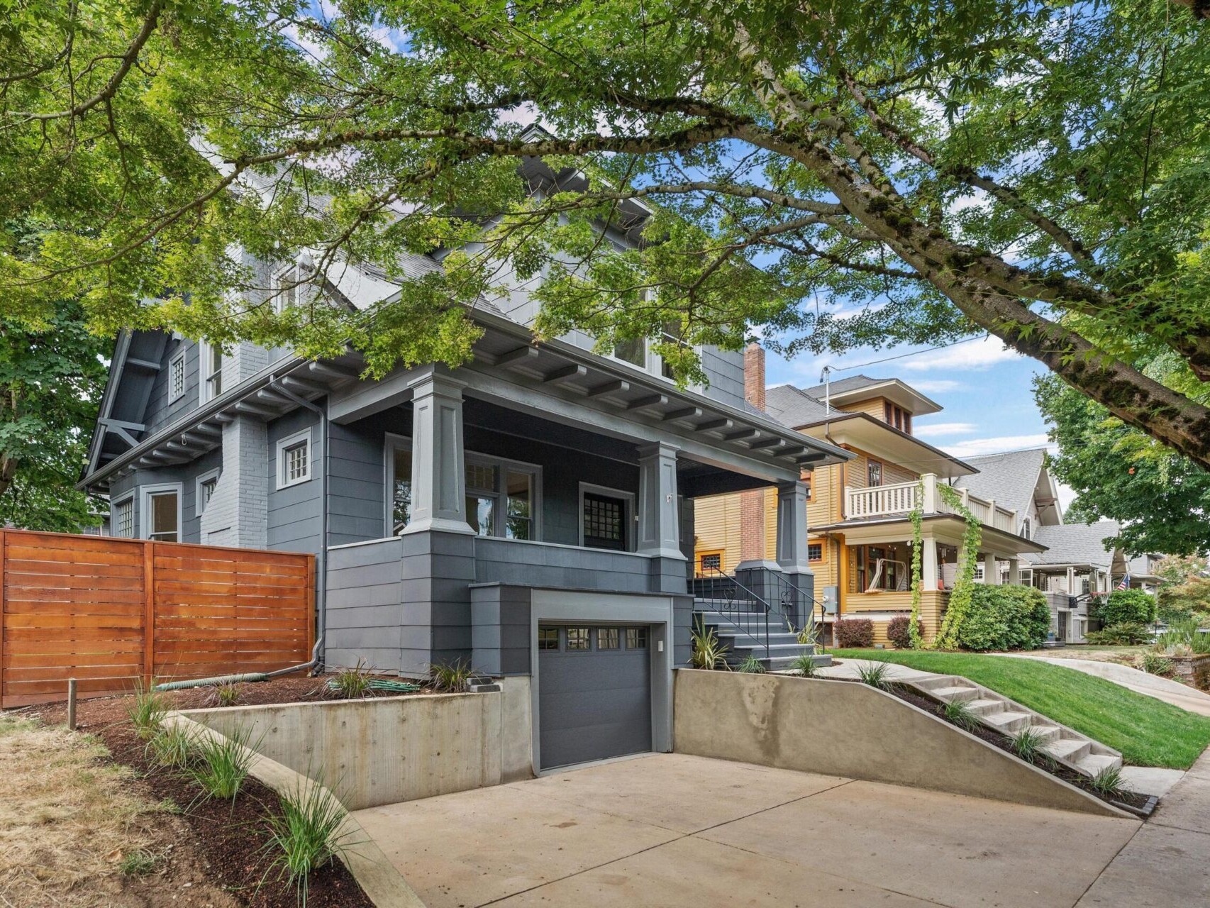 A gray two-story house with a porch and large windows, nestled among Portland, Oregon's lush trees. The front features a driveway leading to a garage, reflecting the area's charm. The neighborhood boasts similar houses and a well-maintained sidewalk typical of this vibrant city.