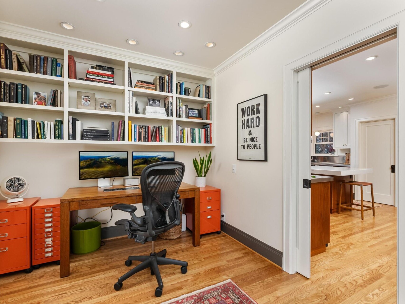 Home office featuring a wooden desk, dual monitors, and a black chair. Red drawers and a green plant nestled beneath the desk. White shelves brimming with books and decor above, alongside a motivational poster. Ideal for any top real estate agent in Portland Oregon, it sits adjacent to a kitchen space with wooden flooring.