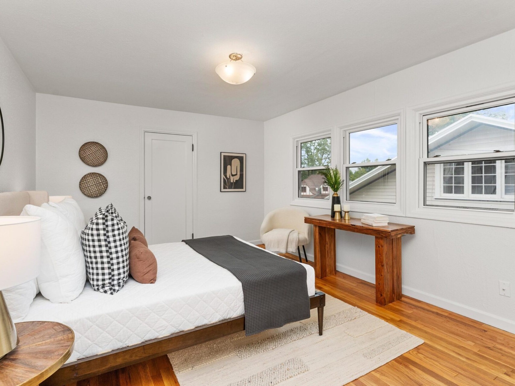 A cozy bedroom with a neatly made bed, featuring white and checkered pillows. A wooden side table and a matching desk add warmth. Three windows provide ample natural light, and wall decor includes a round mirror and woven art pieces.